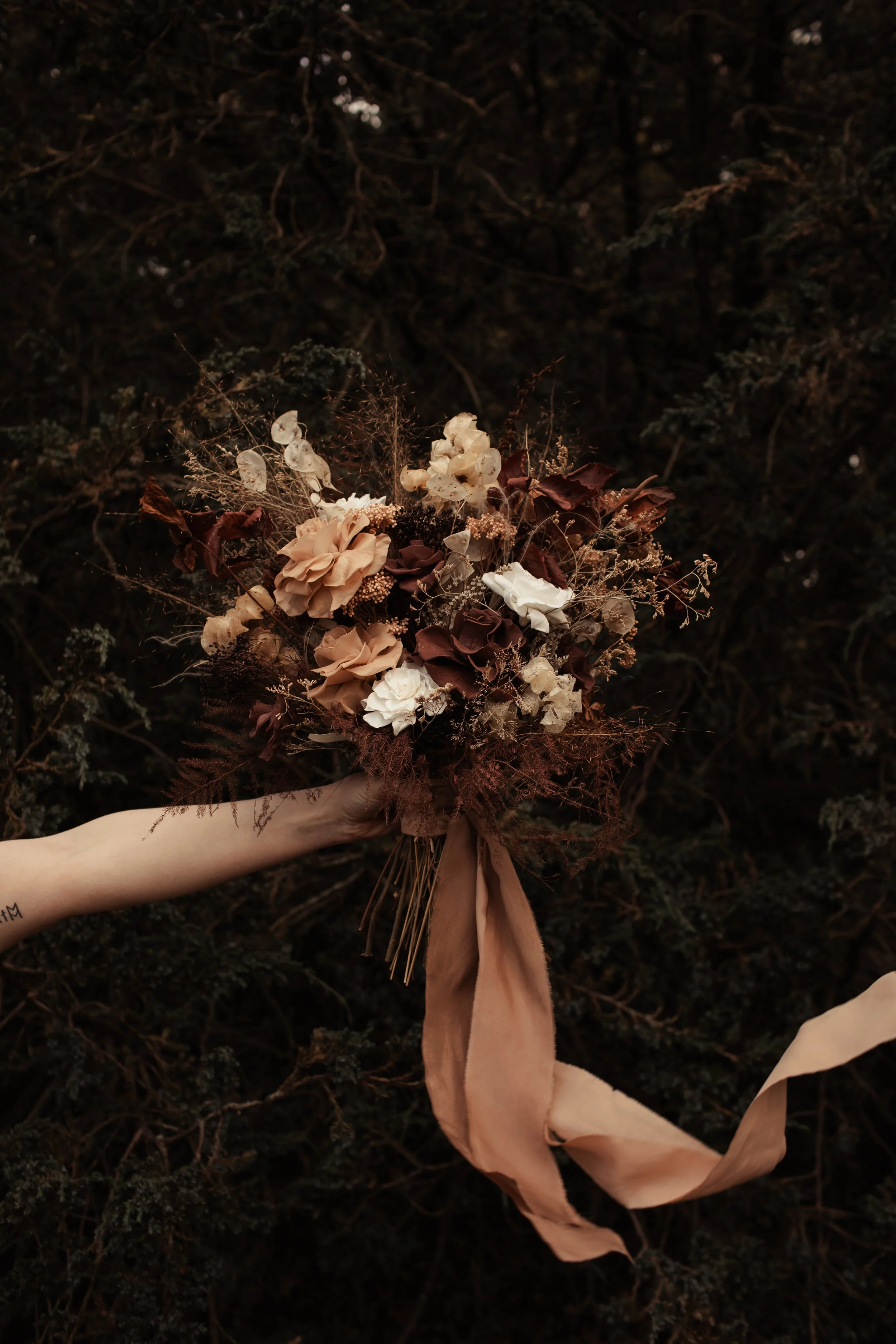 A hand holding a bouquet of dried flowers with brown, beige, and white blossoms and long beige ribbon against a dark, bushy background.