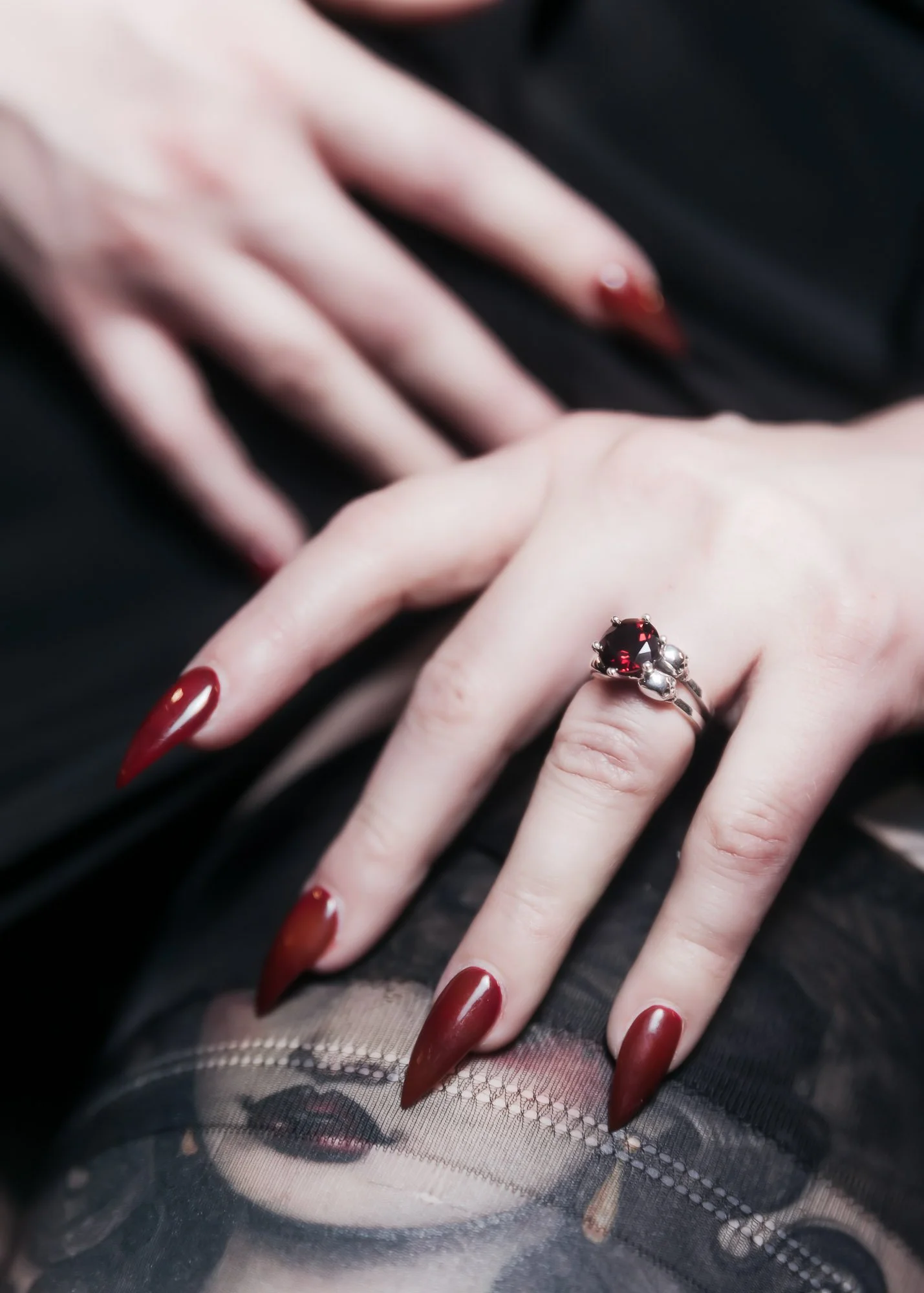 Close-up of a hand with long, pointed, red nails wearing a ring with a large red gemstone. The hand is resting on a black fabric surface, and underneath is a sheer fabric with a printing of a woman's face.