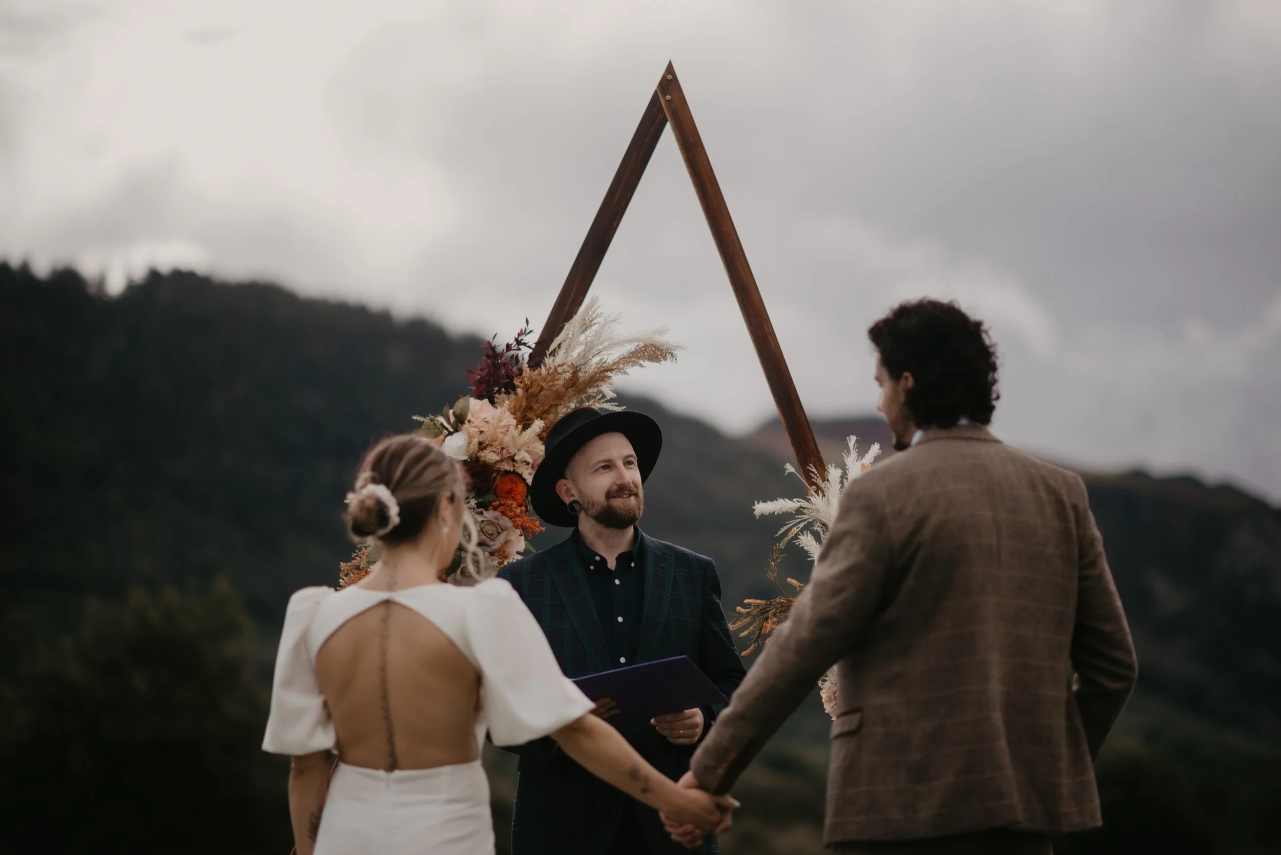 A wedding ceremony outdoors with a couple holding hands and standing in front of an officiant, in front of a mountain landscape and a wooden triangular arch with floral decorations.