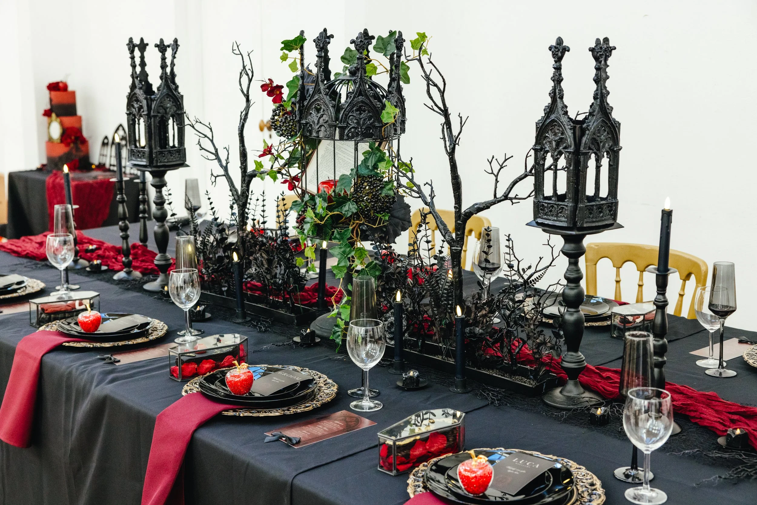 A Halloween-themed dining table setup with black and red decorations, lanterns, candles, tall black candlesticks, black and gold plates, and apple-shaped decorations, with a black and red table runner and green foliage accents.