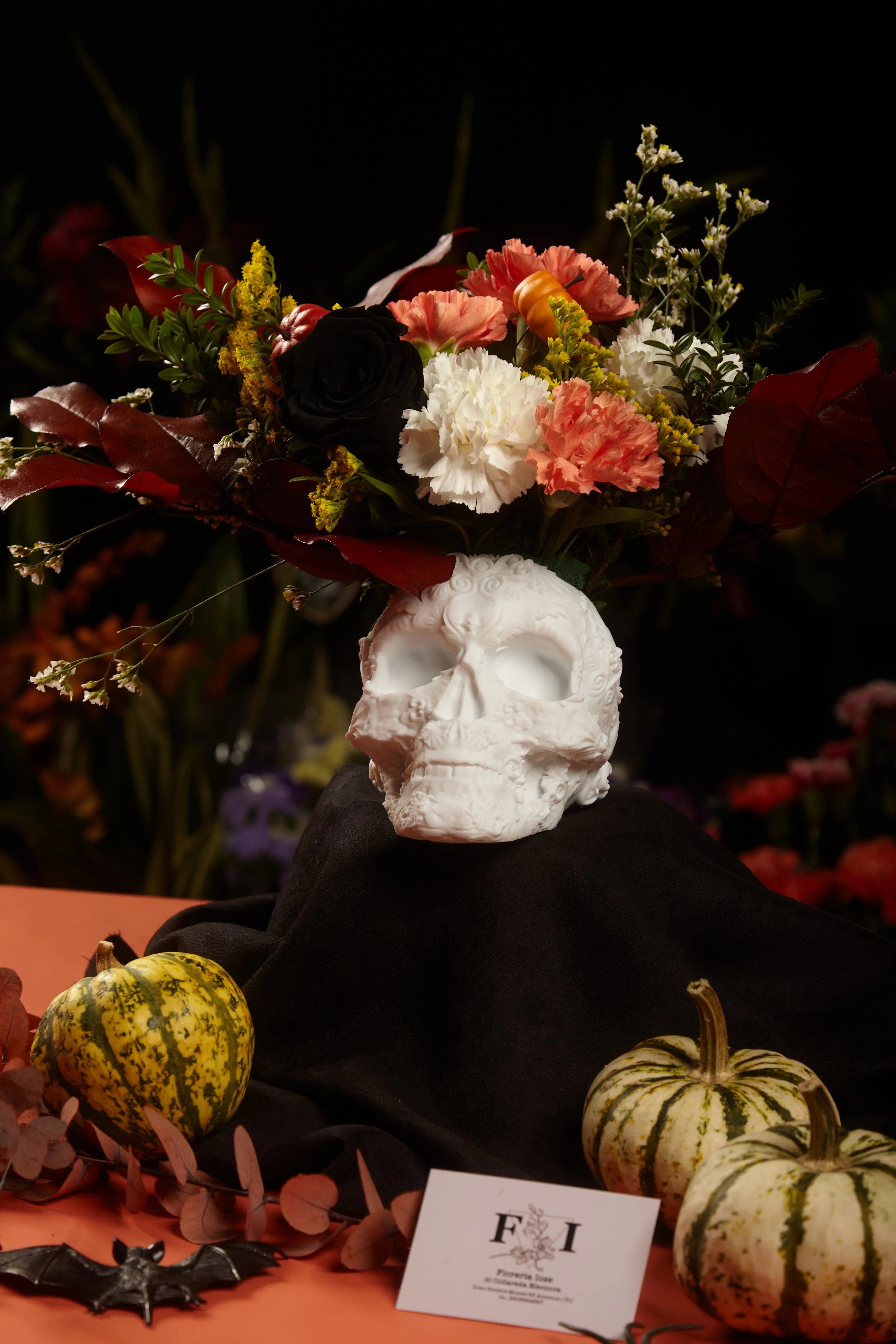 Arrangement of flowers in a skull-shaped vase, with decorative pumpkins and Halloween-themed decorations on a table, likely for a fall or Halloween display.