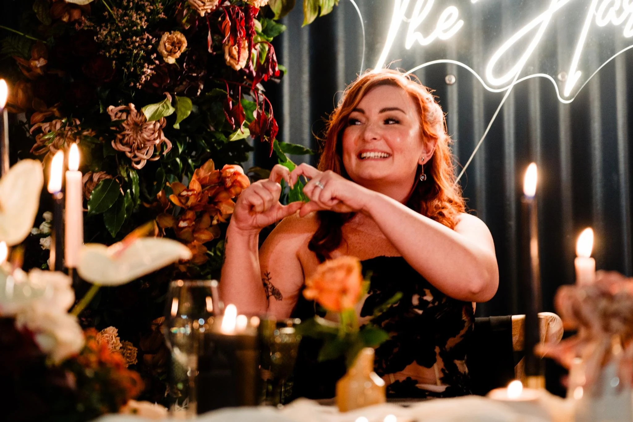 A woman with red hair smiling and making a heart shape with her hands at a decorated party table with candles, flowers, and a neon sign in the background.
