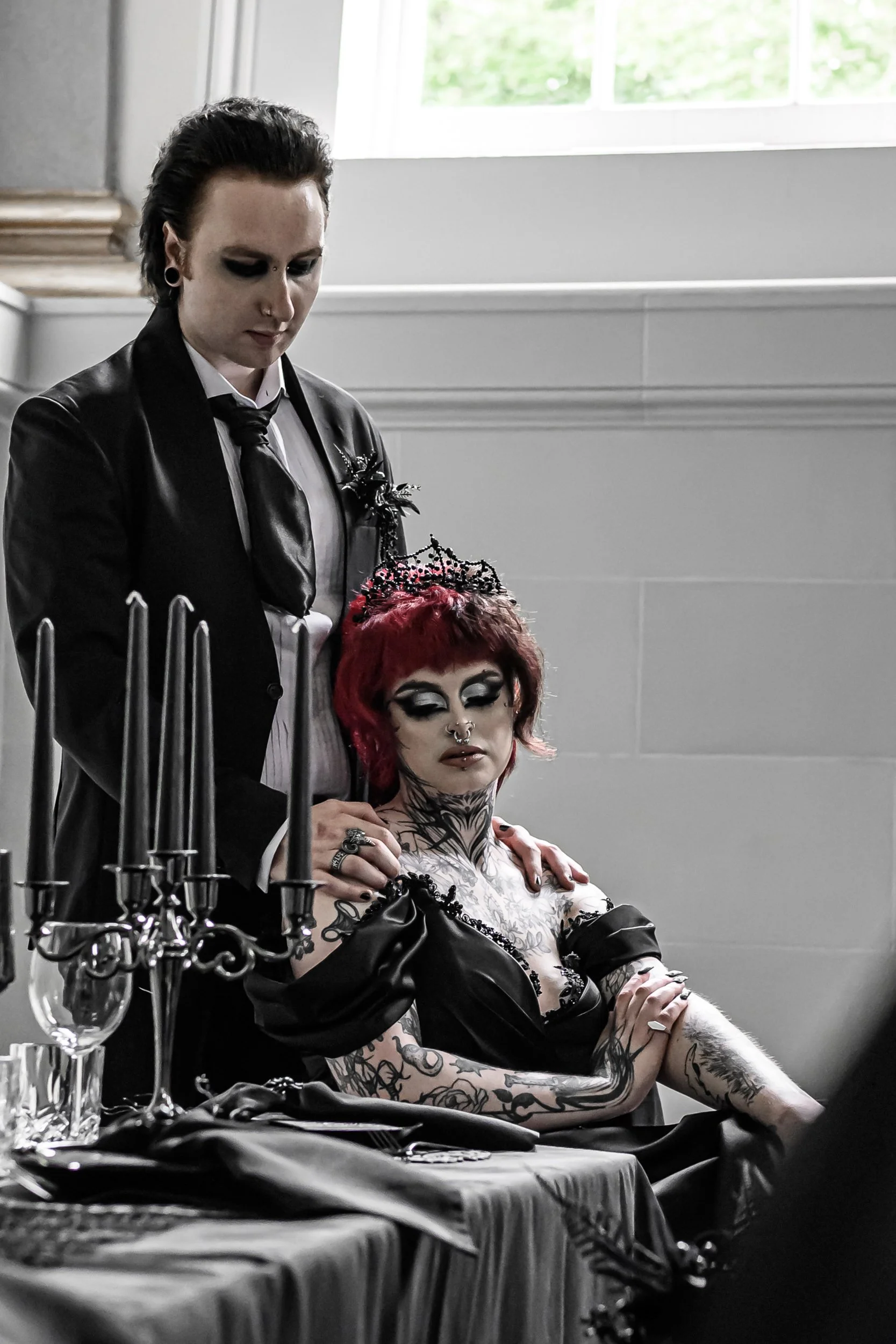 A person with vibrant red hair, heavy makeup, tattoos, and gothic clothing sitting at a table with a black dress, silver jewelry, and a candelabrum, while another person in a black suit and white shirt with a black tie stands beside them, touching th