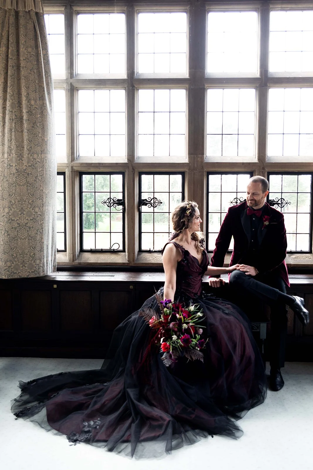 A bride and groom sitting by a large window with multiple panes, the bride holding a bouquet of dark red and purple flowers, both dressed in dark formal attire, with the bride in a gown and the groom in a tuxedo, looking at each other inside a histor