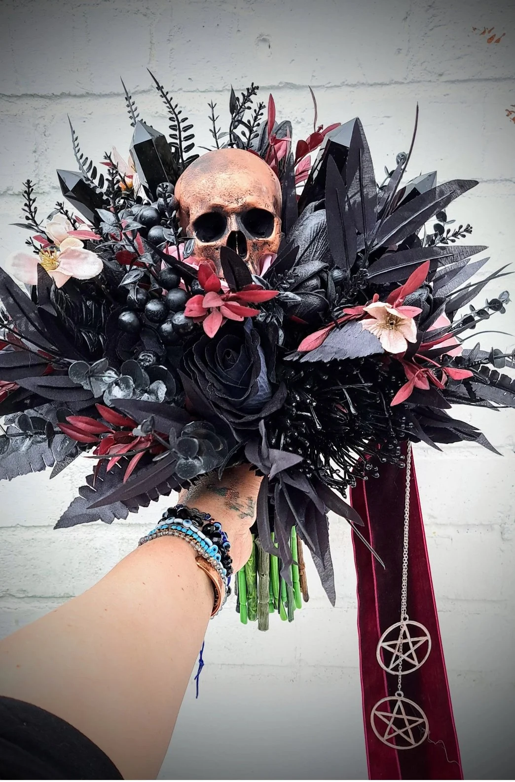 A bouquet of black and red artificial flowers with a fake skull at the center, decorated with black leaves, berries, and pink flowers, held by a person with bracelets, against a plain white wall.