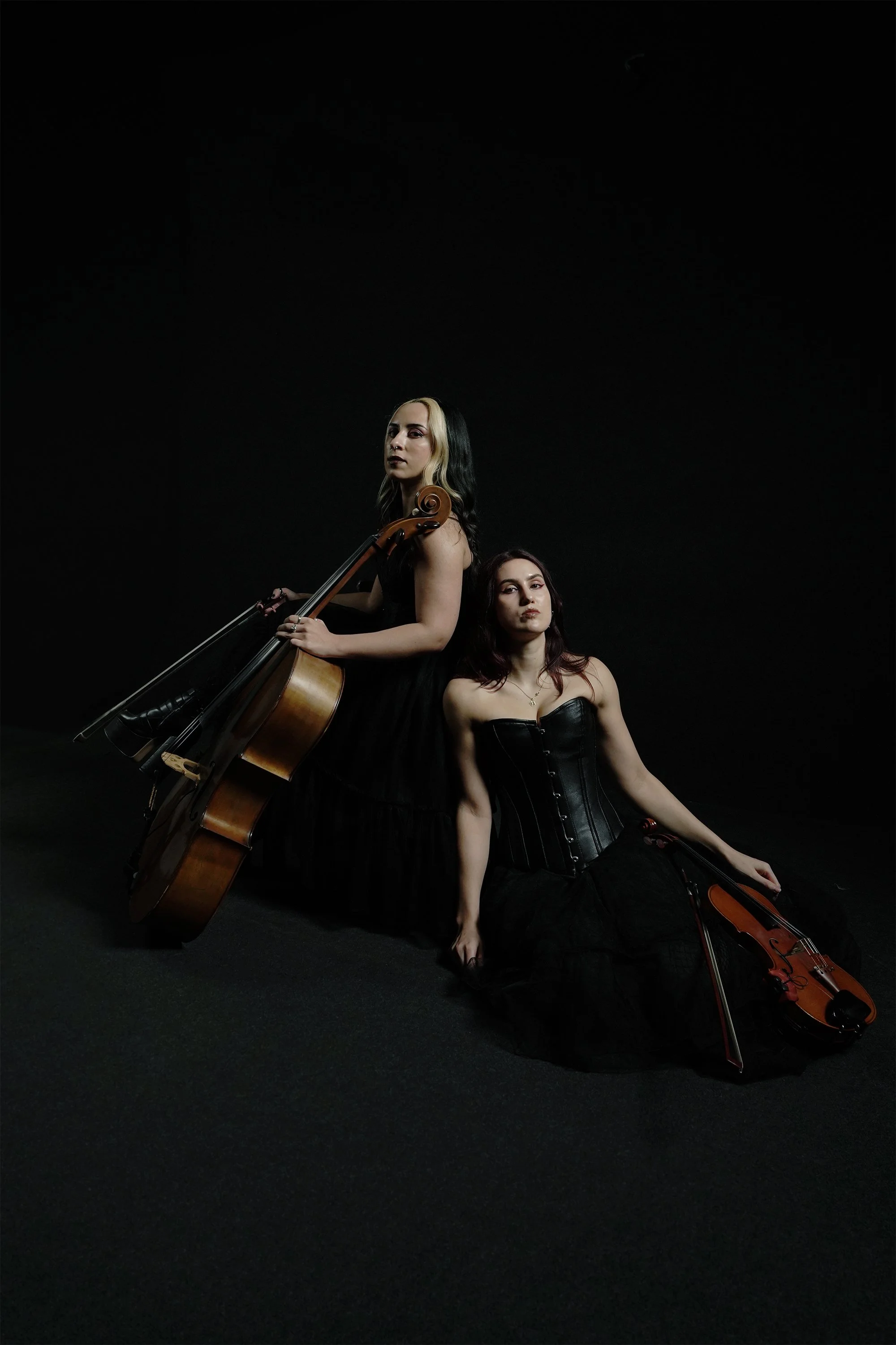 Two women in black dresses with musical instruments, one holding a cello and the other with a violin, sit against a dark background.