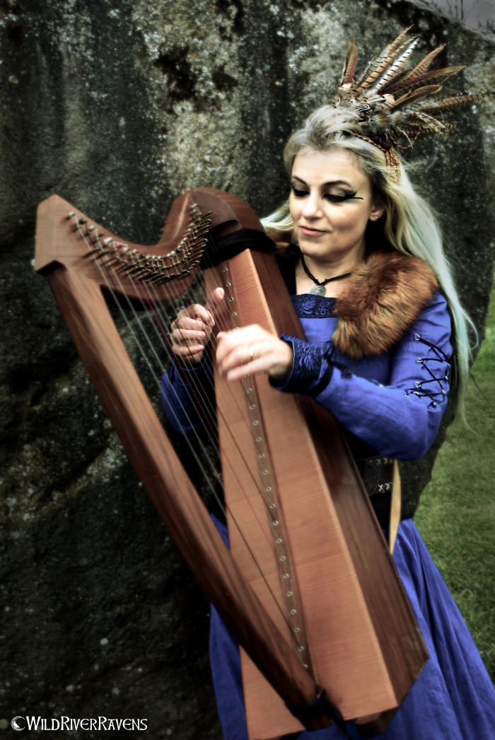A woman dressed in medieval or fantasy costume, wearing a blue dress with fur and feather accessories, playing a harp outdoors near a large stone wall.