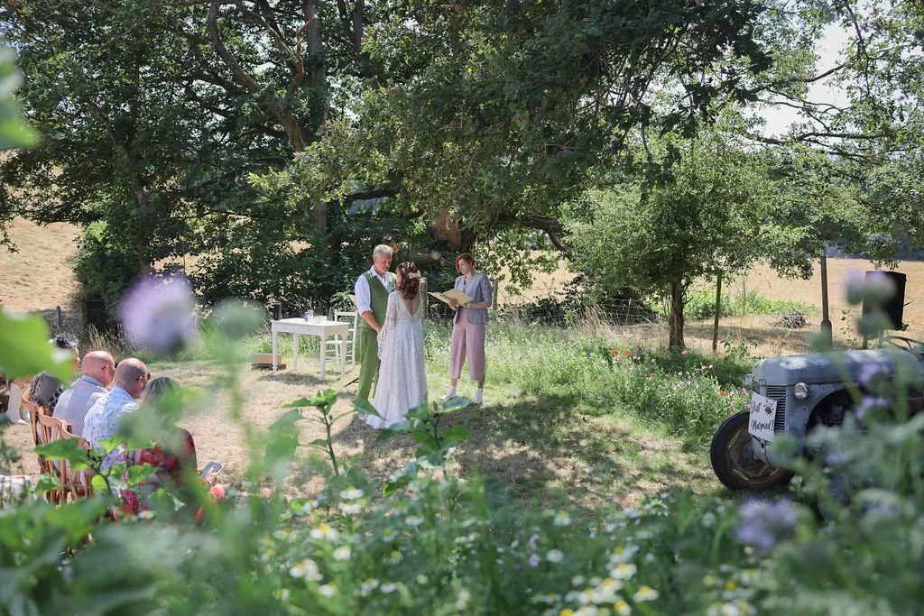 A couple getting married outdoors under a large tree, with an officiant, in front of guests seated on wooden chairs, surrounded by greenery and flowers.