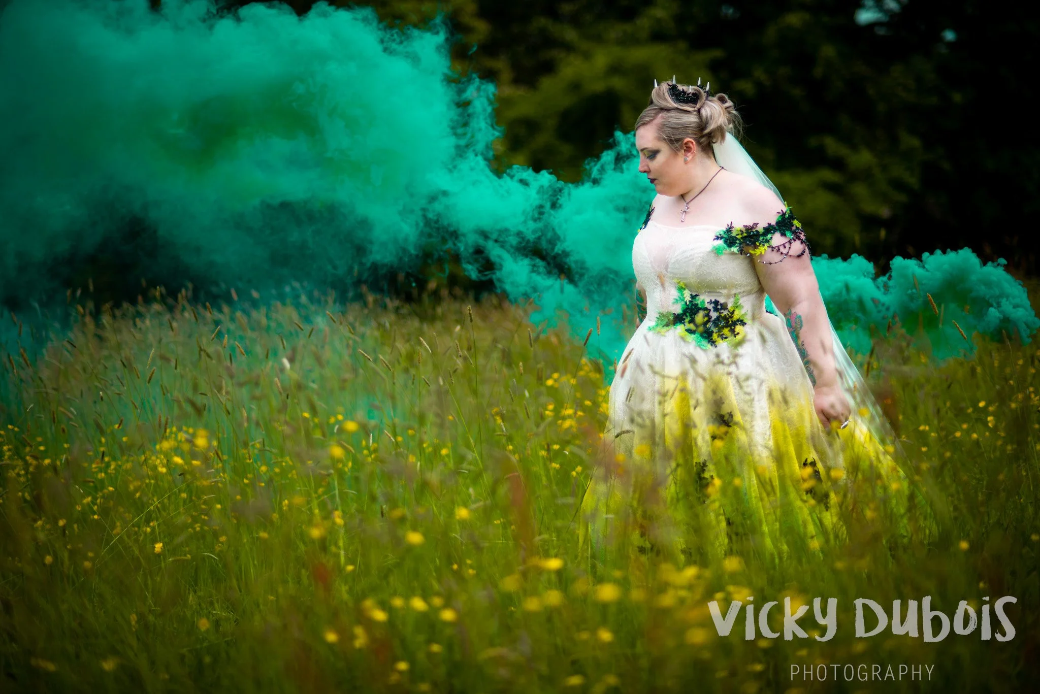 A woman in a cream-colored dress with black and green floral details stands in a grassy field with yellow flowers. She holds a green smoke bomb emitting green smoke, with a dark forest background. Her hair is styled up with a crown and veil.