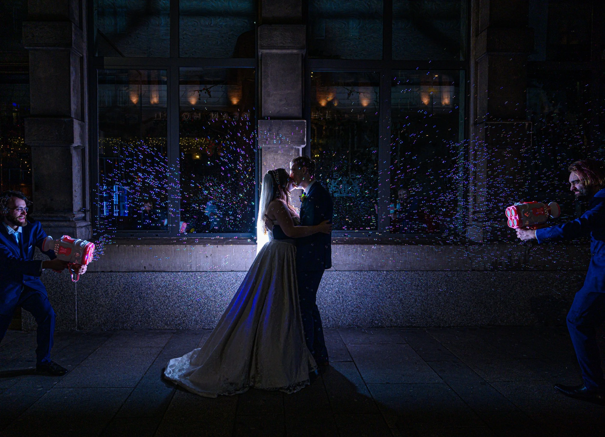 A bride and groom kiss in the center of a nighttime scene, backlit with a bright light behind them creating a silhouette effect, with two men shooting confetti cannons on either side of them and colorful confetti in the air.