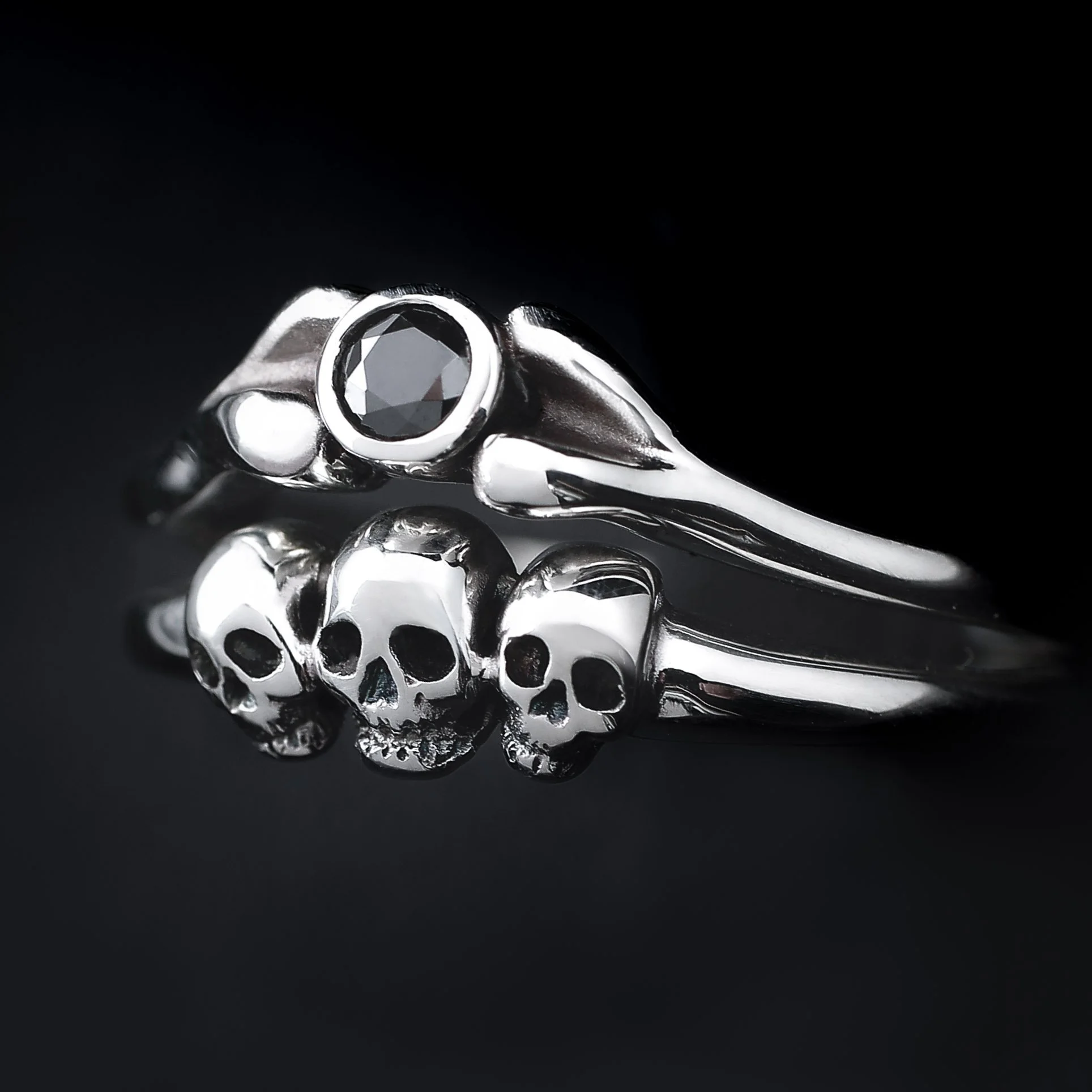 Silver ring with a black gemstone on top and three small skull designs on the band, against a black background.