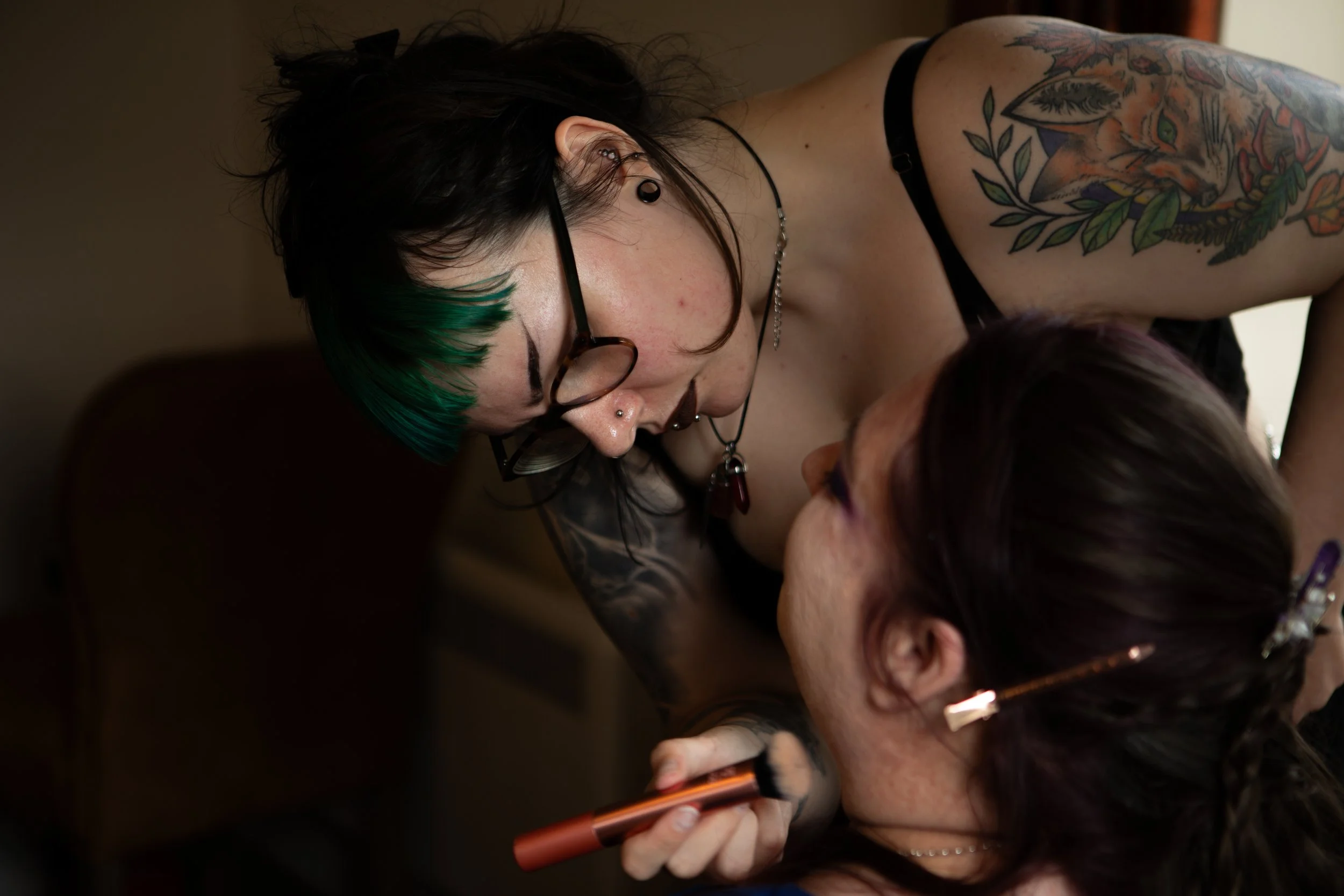 A woman with green-tipped black hair and tattoos applying makeup to another woman with red hair, who is lying down.