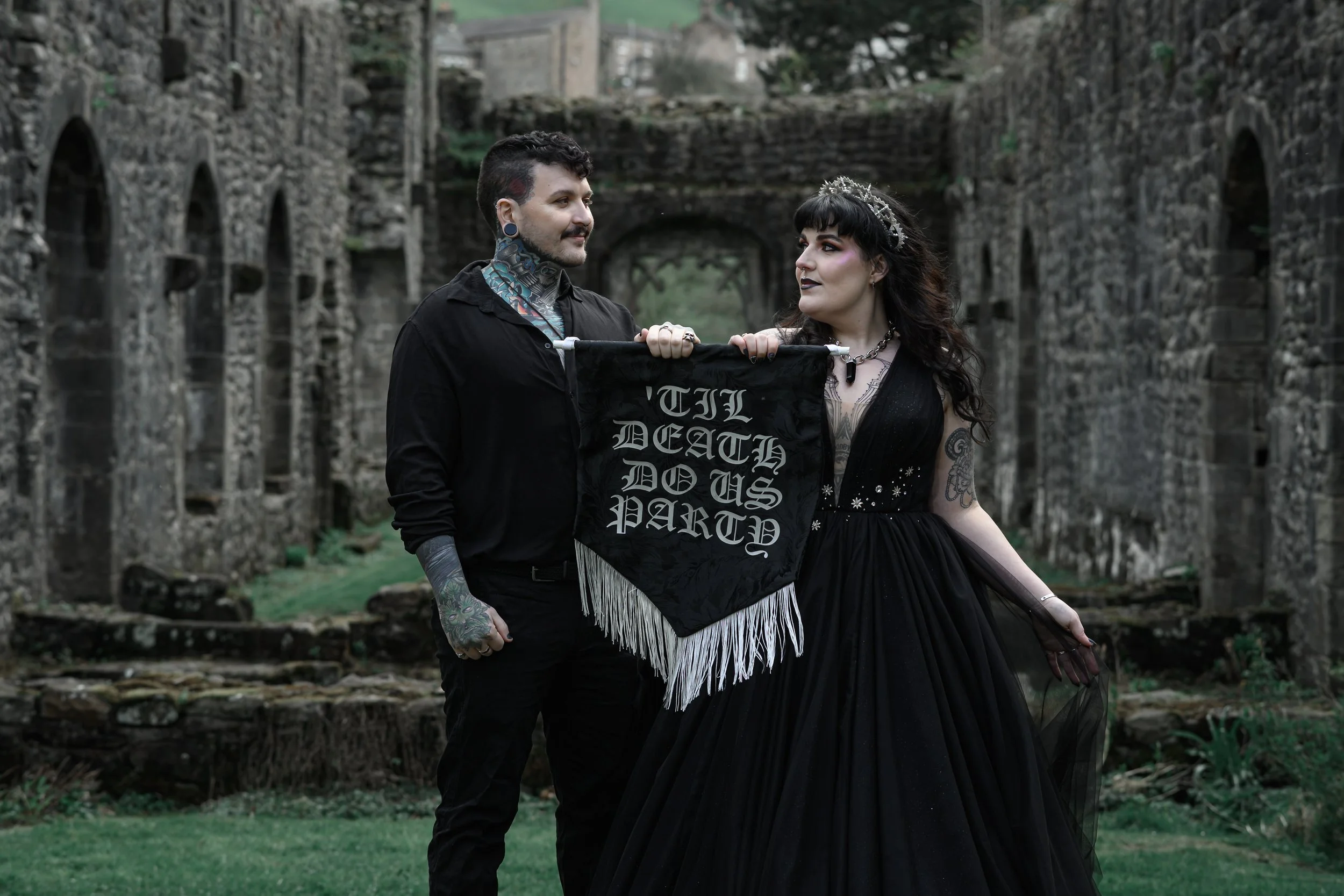 A goth couple stands outdoors holding a banner that says, 'Till Death Do Us Part,' with ruins and greenery in the background.