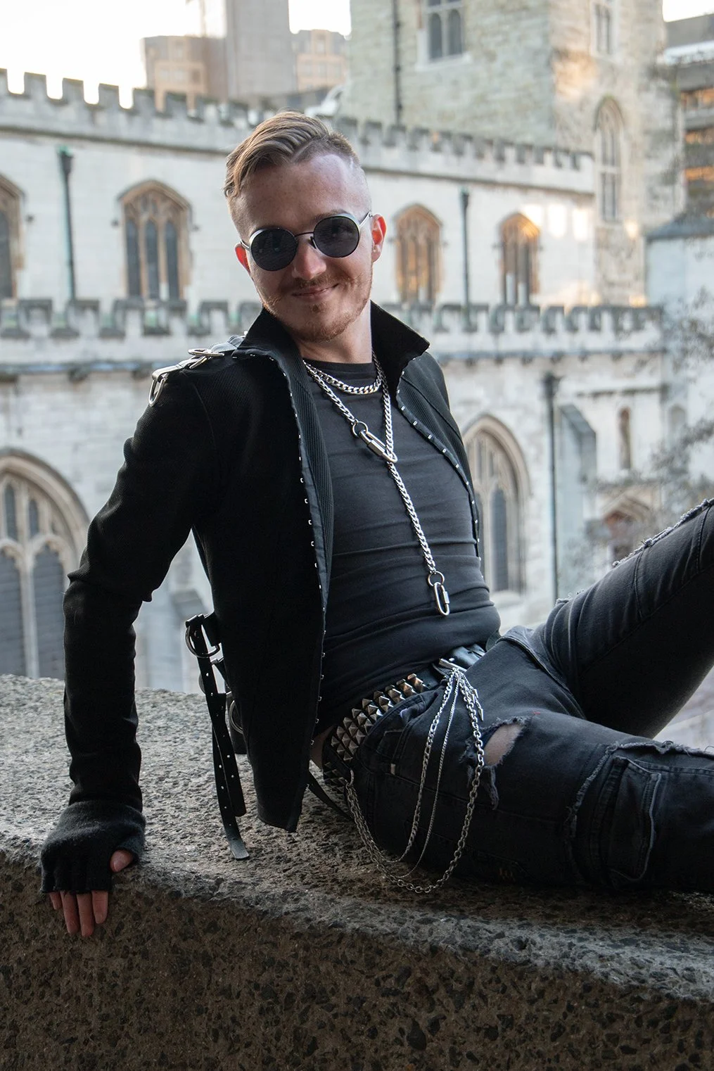 A young man with styled hair and sunglasses sitting on a stone ledge in front of a historic stone building, wearing black clothing, chains, and accessories.