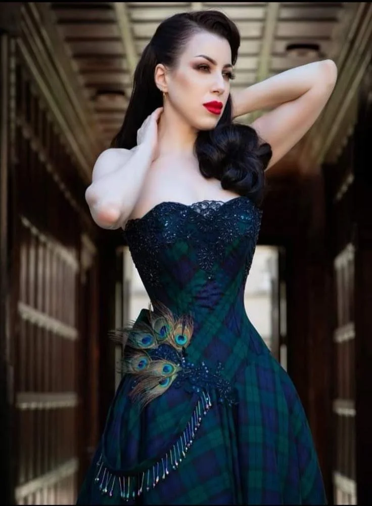 A woman with dark hair and red lipstick wearing a strapless dress with peacock feather details in an indoor hallway.