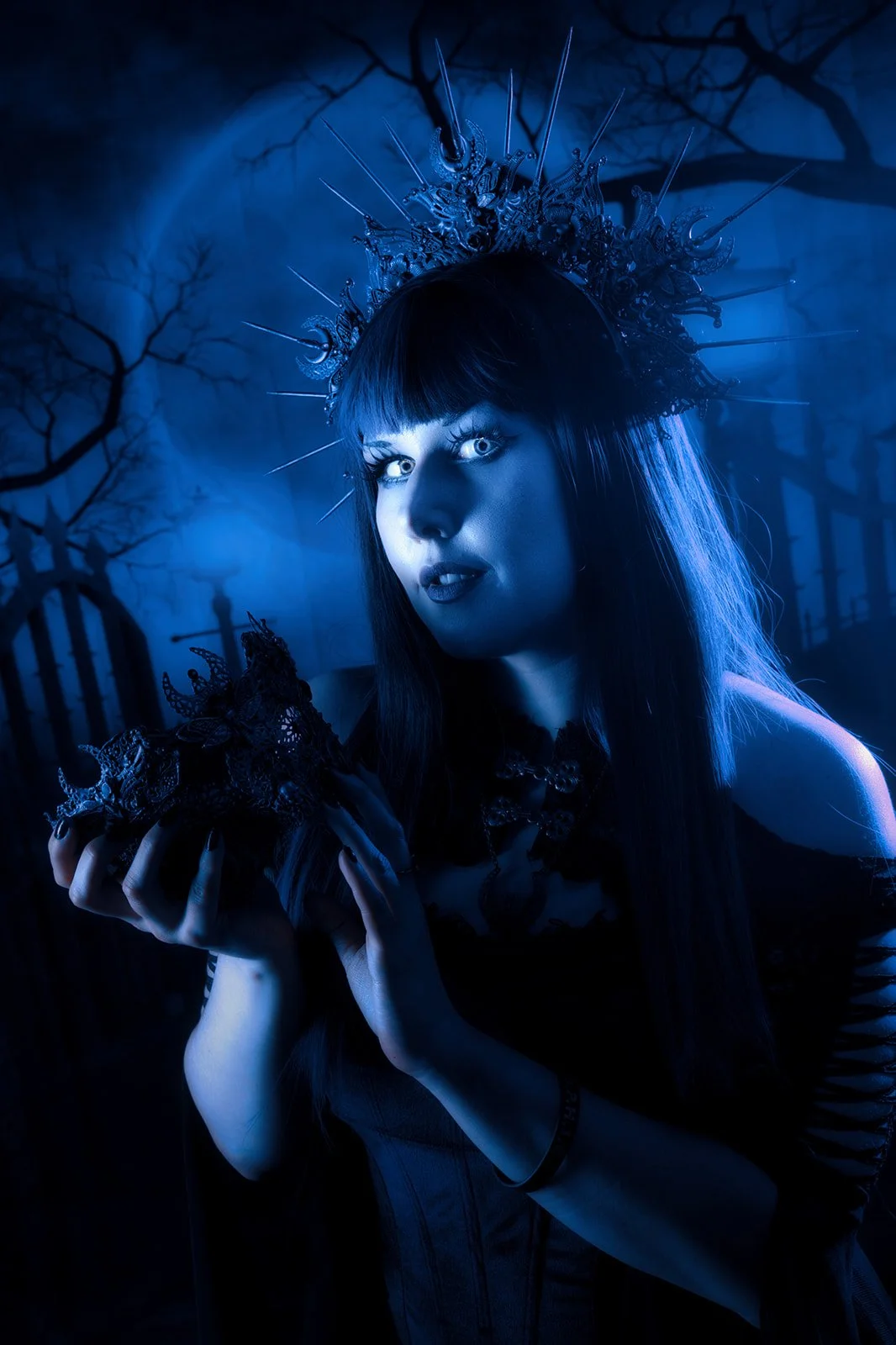 A woman with dark hair and a crown made of spikes, holding a scepter or staff, standing outdoors in a dark, eerie forest with leafless trees in the background, illuminated with bluish lighting.