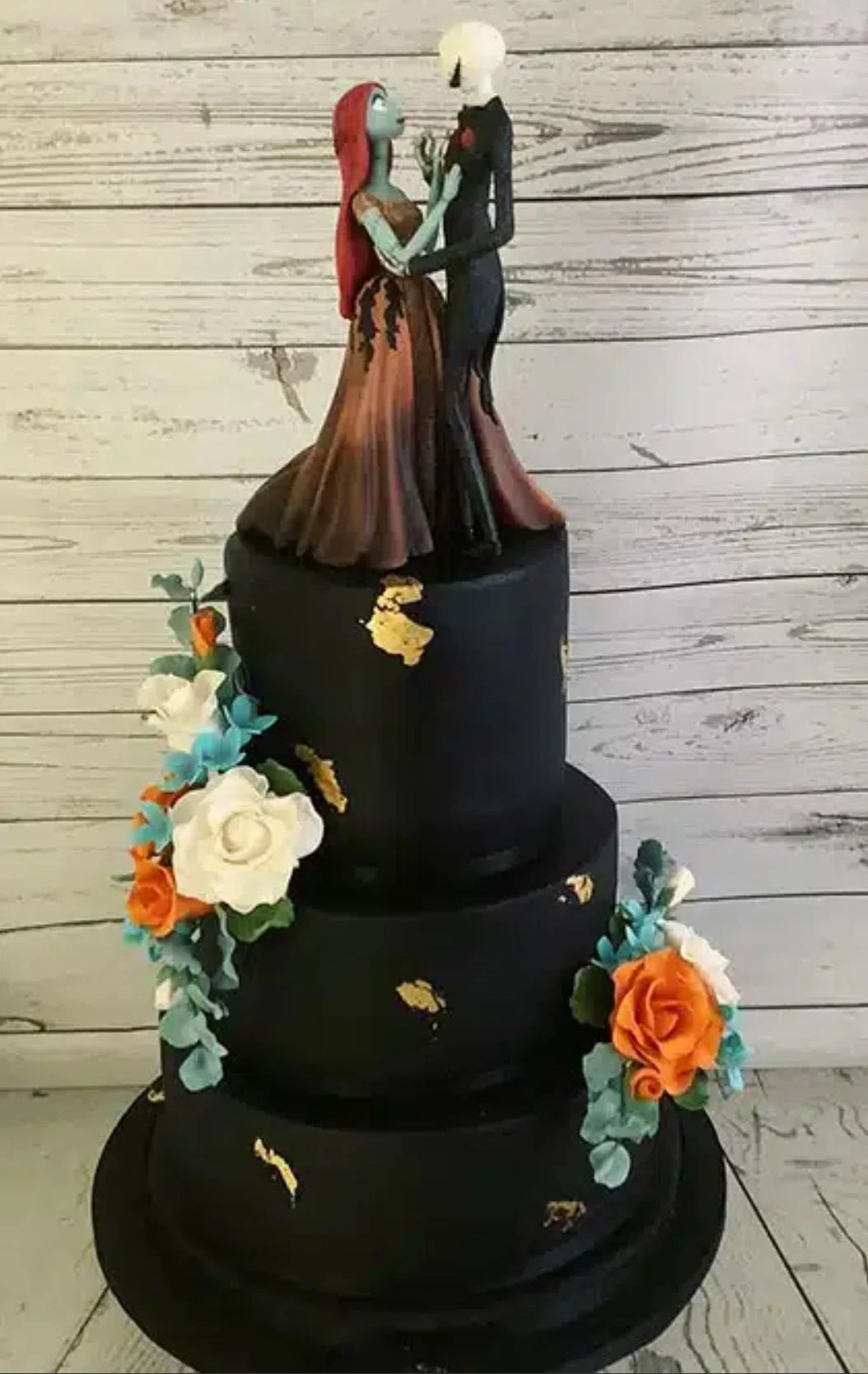A three-tier black wedding cake with gold leaf accents and colorful flower decorations, topped with a figurine of a bride with red hair in a brown gown and a groom with blonde hair in a black tuxedo, standing on a wooden background.