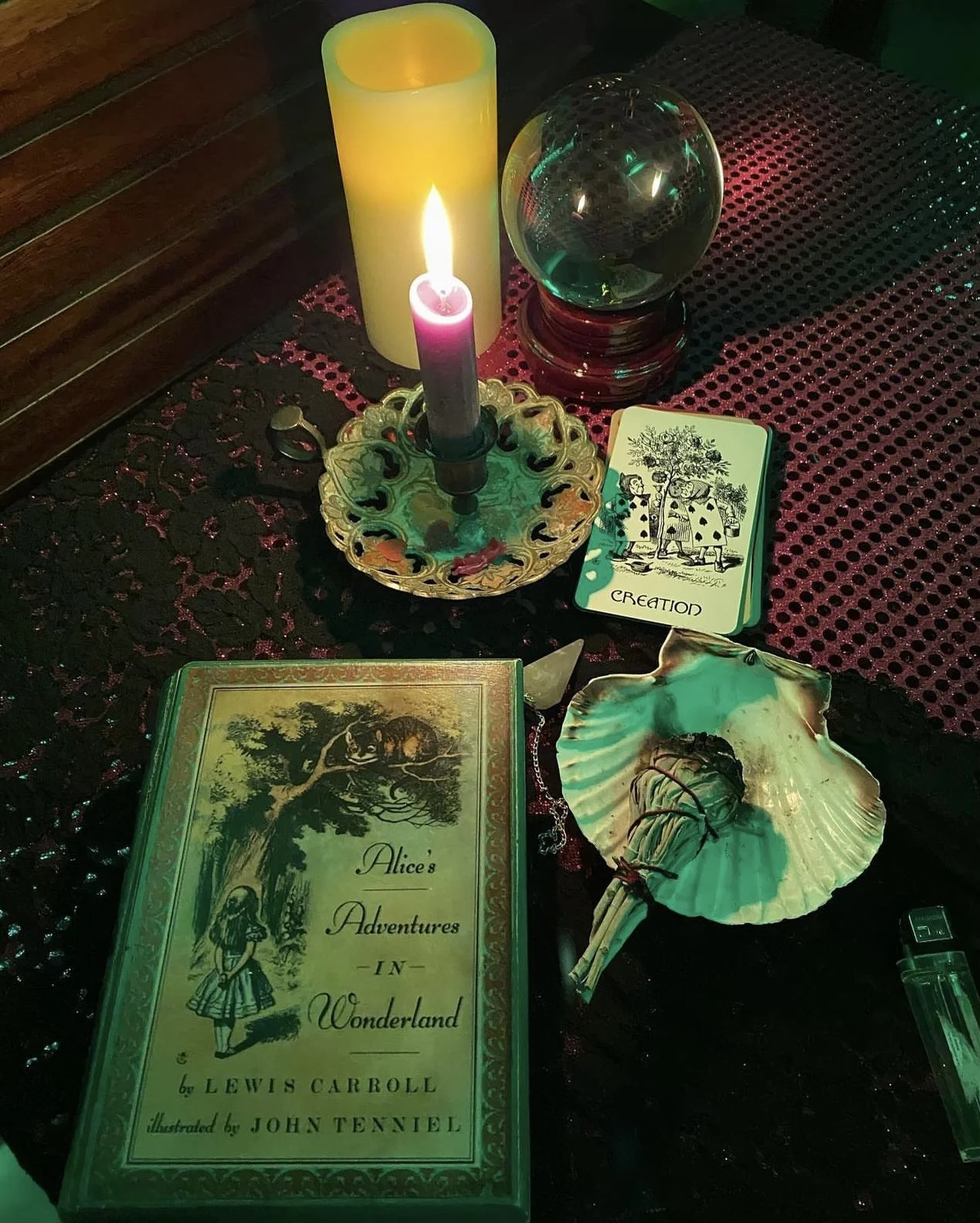 A table setup with a vintage book titled 'Alice's Adventures in Wonderland', a tarot card deck, a seashell holding a small object, a crystal ball, a candle, and a lit candle in a holder, all on a decorative dark table cover.