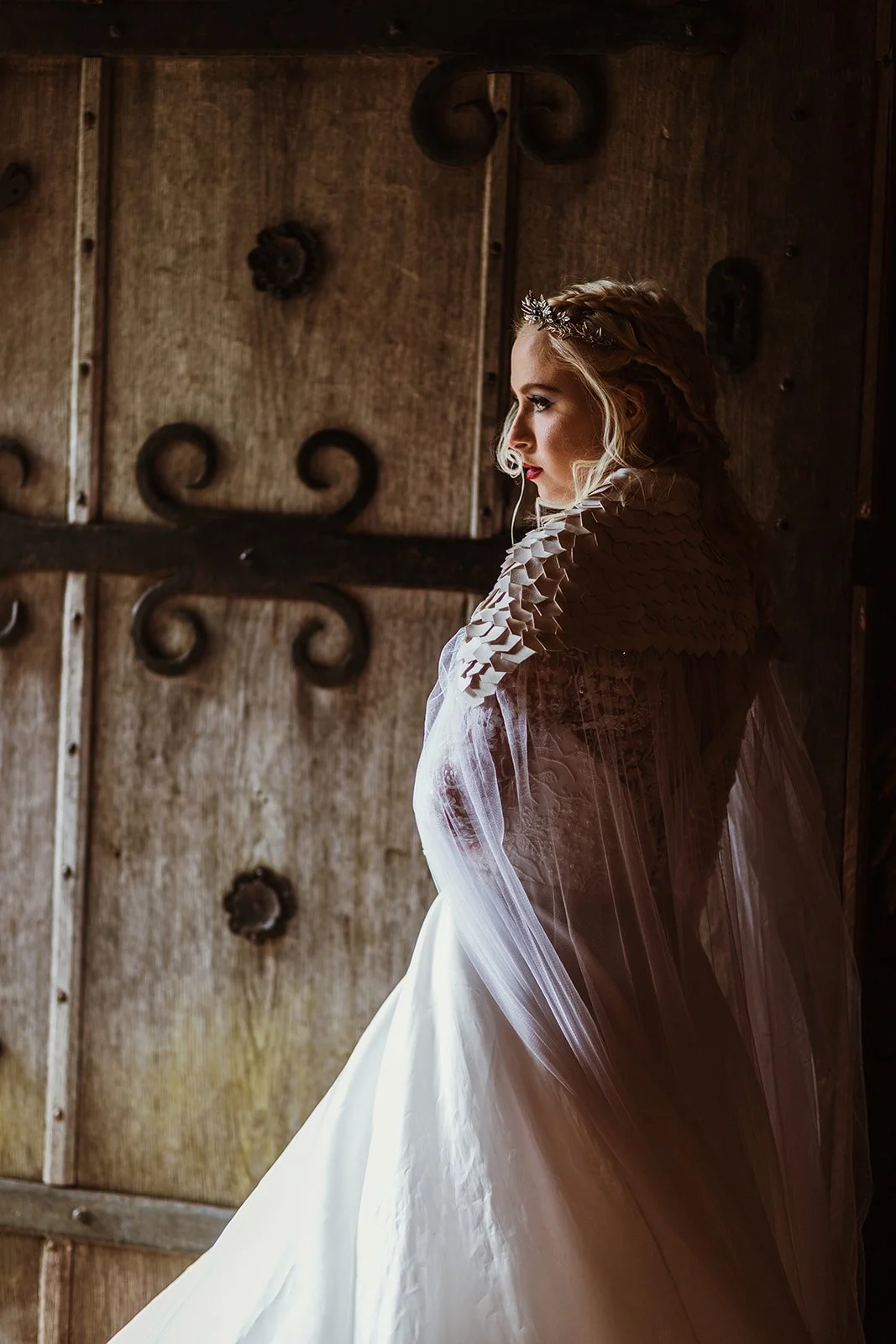 A bride with styled hair, wearing a wedding dress and veil, stands in profile by a rustic wooden door with metal accents.