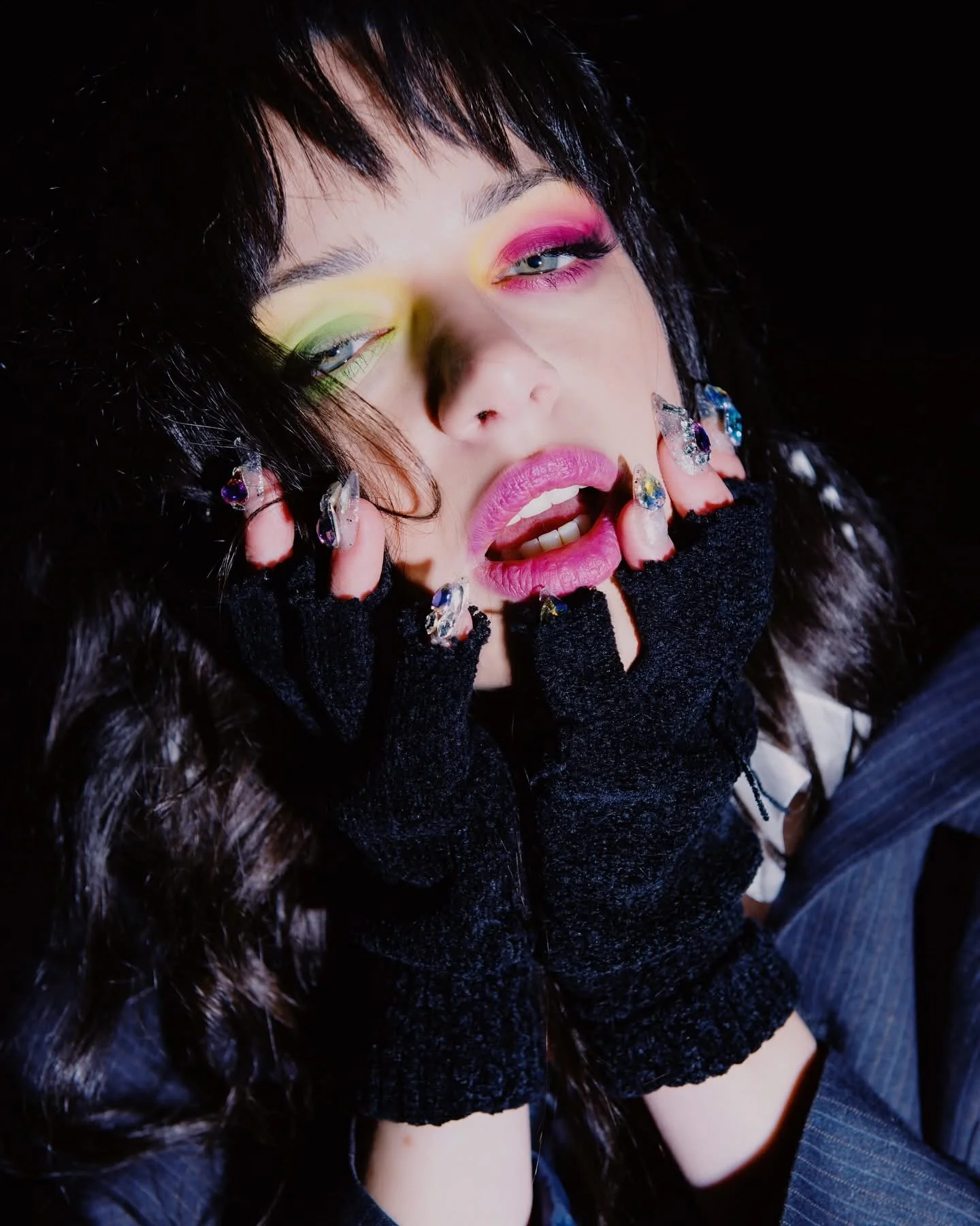 A person with colorful eye makeup, pink lipstick, and decorated nails, wearing black gloves with jewelry, touching their face with a dramatic expression.