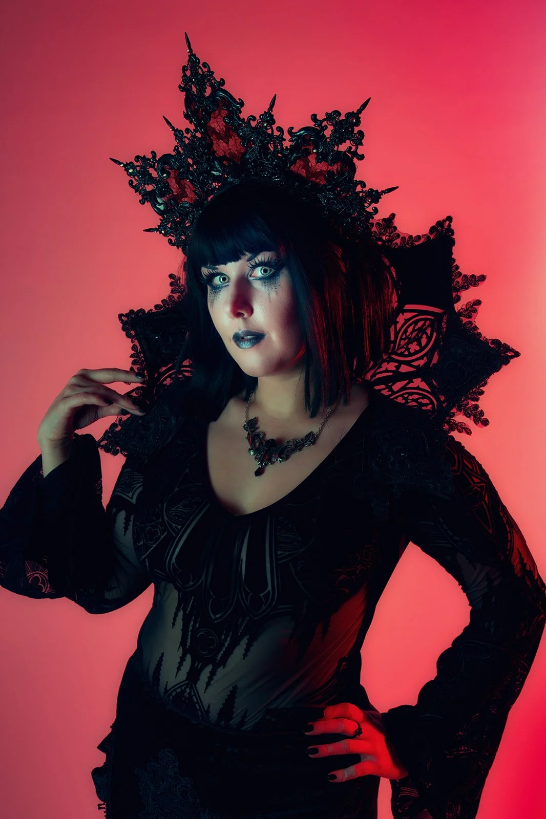 Woman in gothic costume with elaborate black lace headpiece and dark makeup against pink background.