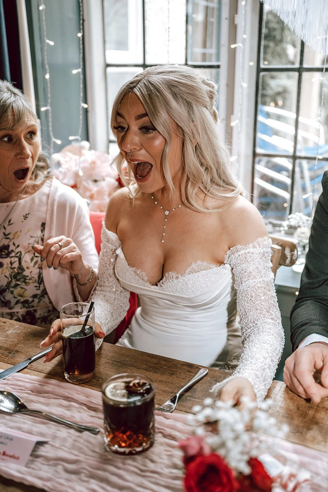 A woman in a wedding dress appears surprised or excited at a celebration dinner, sitting at a table with drinks, with two other women, and a window in the background.