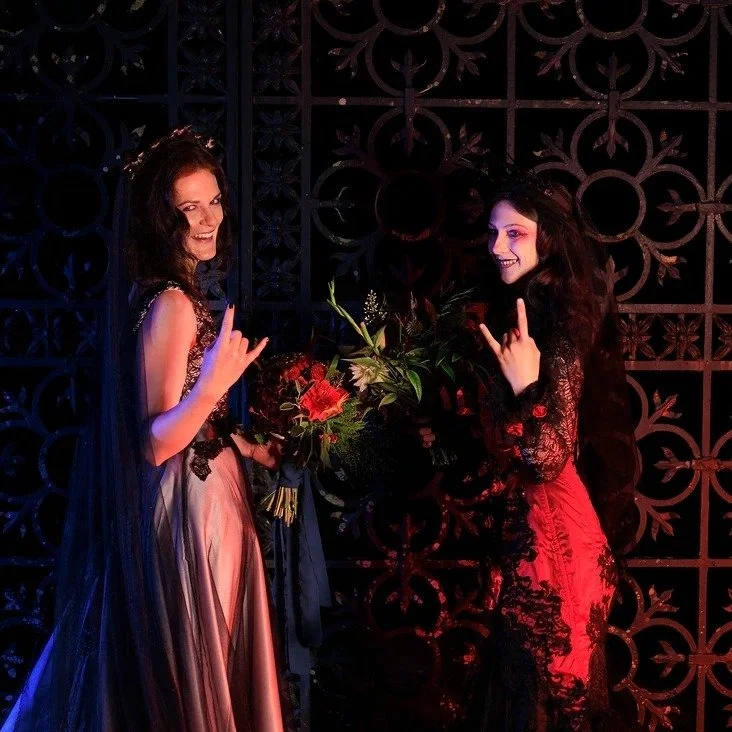 Two women in evening gowns standing on stage, one holding a bouquet of flowers, both smiling and making the 'rock on' hand gesture against a decorative black metal background.