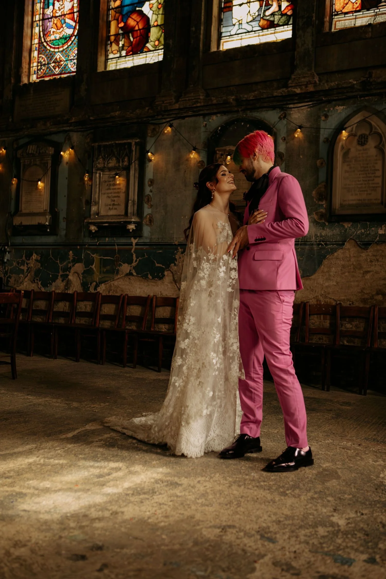 A bride and groom sharing a romantic moment inside a church with stained glass windows and vintage decor, the bride in a lace wedding dress and the groom in a pink suit with pink and green hair.