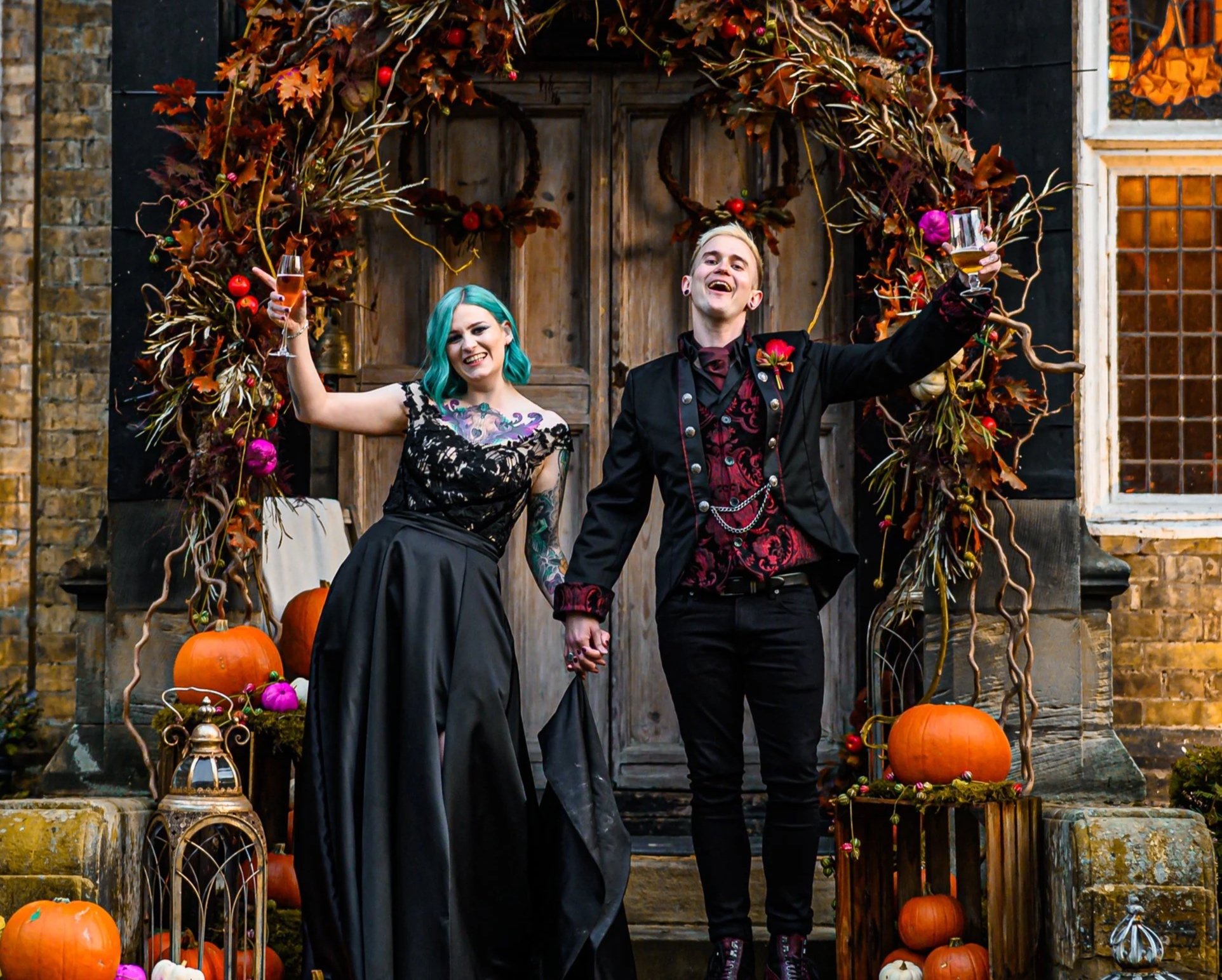 A happy couple dressed in gothic style attire celebrating Halloween on a decorated porch with pumpkins, wreaths, and autumn leaves, holding glasses of wine.