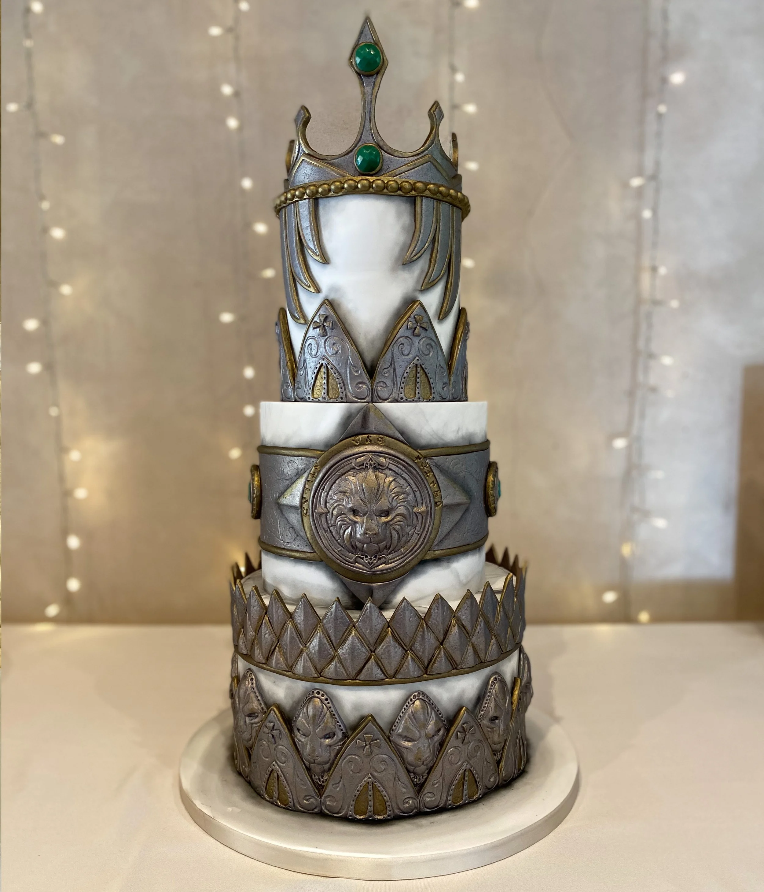 Decorative cake with silver, gold, and white details, featuring a crown top and a lion emblem, displayed on a round white base.