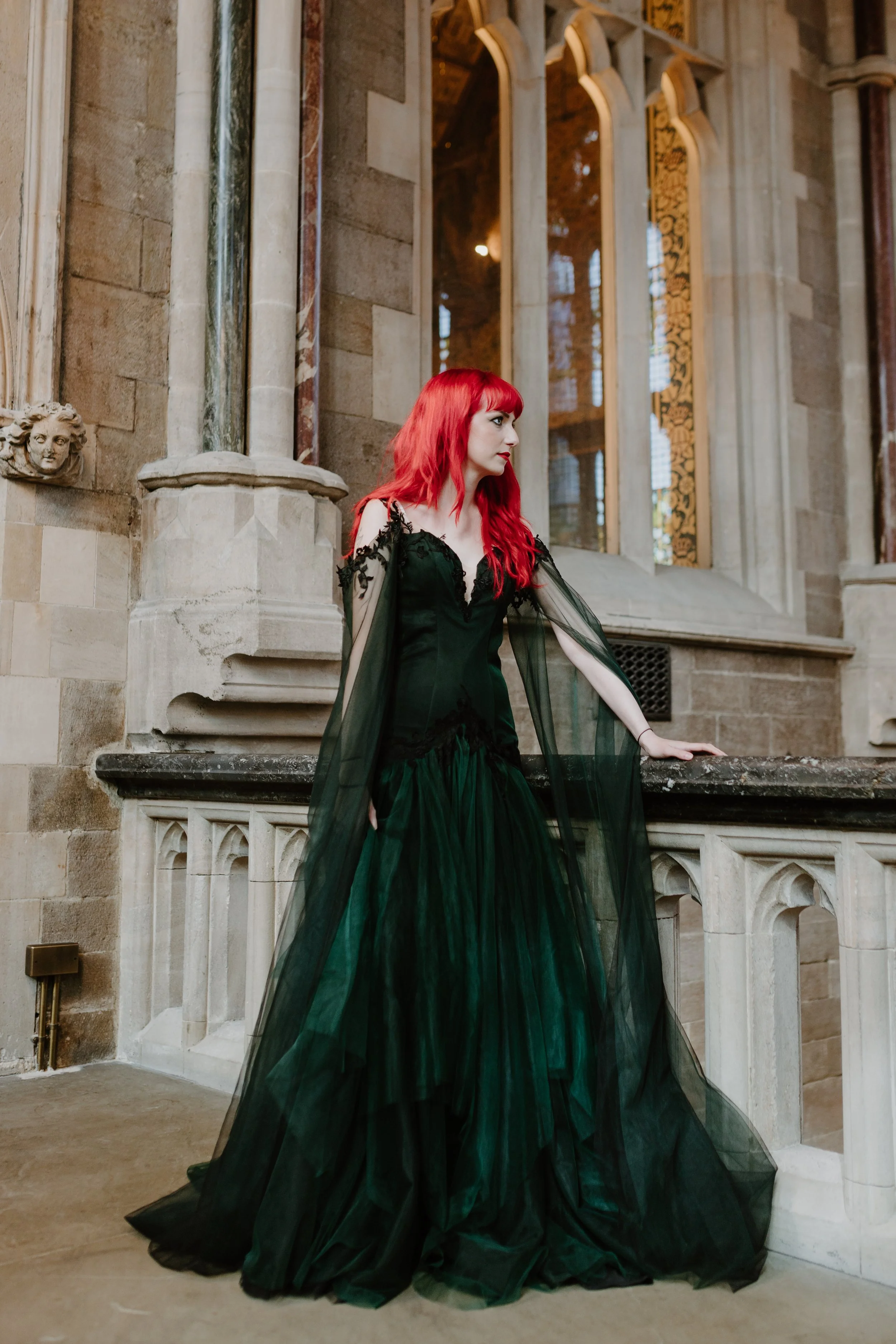 A woman with red hair in a black gown with sheer sleeves stands inside a historic stone building with large windows and ornate architectural details.
