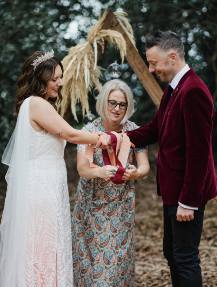 A wedding ceremony outdoors with a bride and groom shaking hands, officiant present, decorated with dried pampas grass and hanging glass orbs.