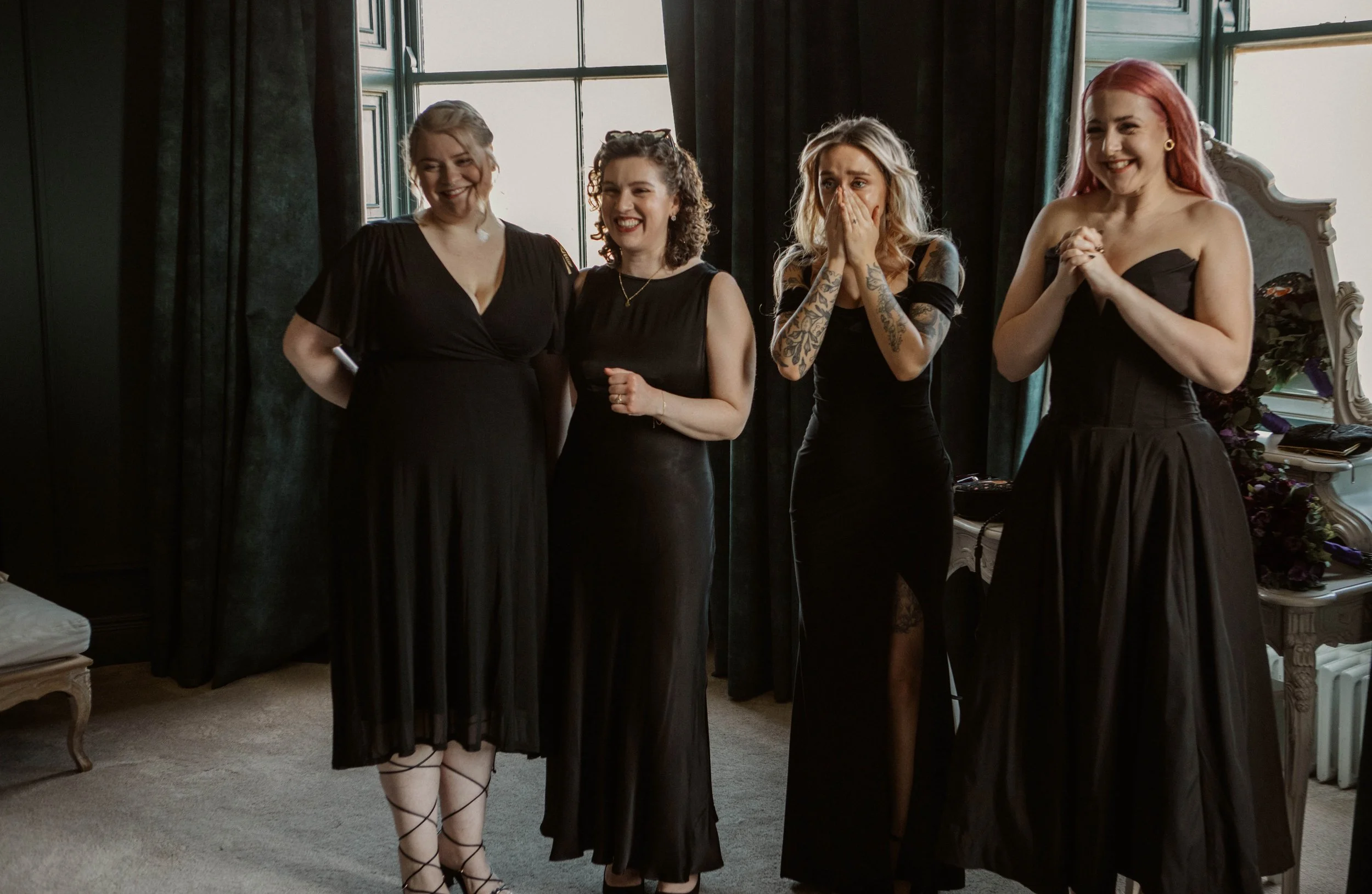 Four women in black dresses standing indoors with large windows in the background. They are smiling, with one woman covering her mouth, appearing to be surprised or emotional.