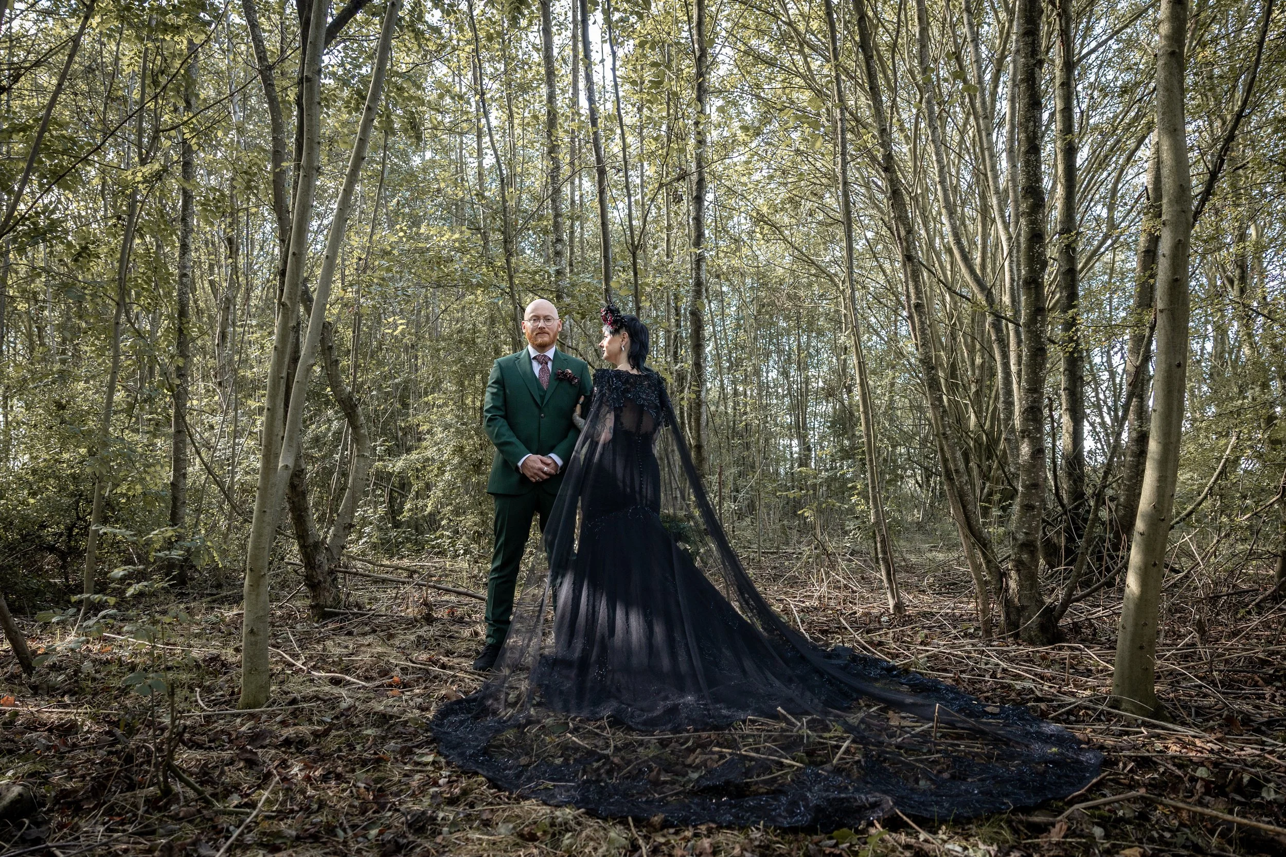 A couple in wedding attire standing in a wooded forest, with the bride wearing a long, black lace dress with a train and the groom in a green suit.