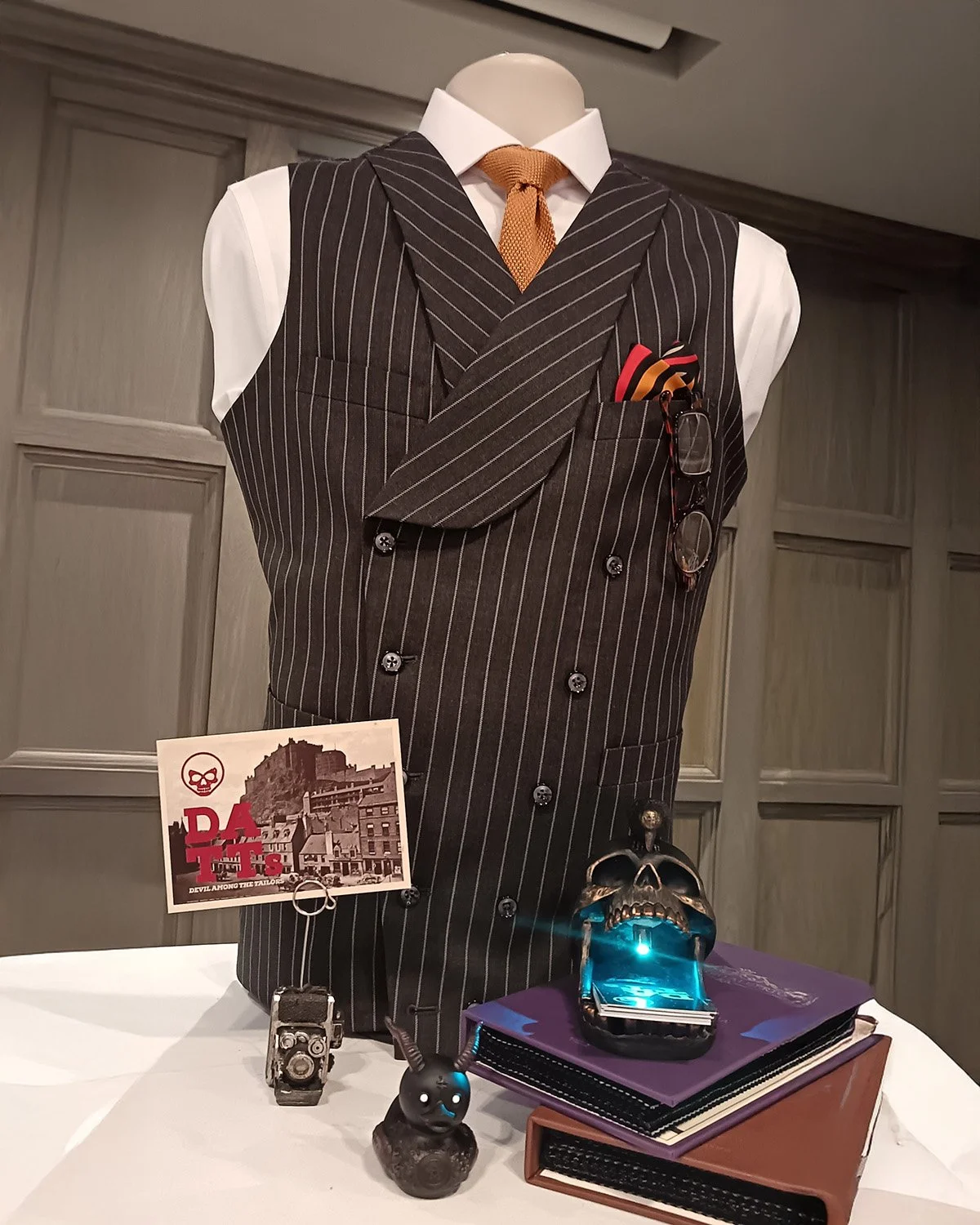 Display of various items including a mannequin dressed in a striped pinstripe vest, white shirt, and an orange tie, with glasses in the pocket. On the table, there is a vintage camera, a small creepy doll, a skull-shaped gadget with a glowing blue li