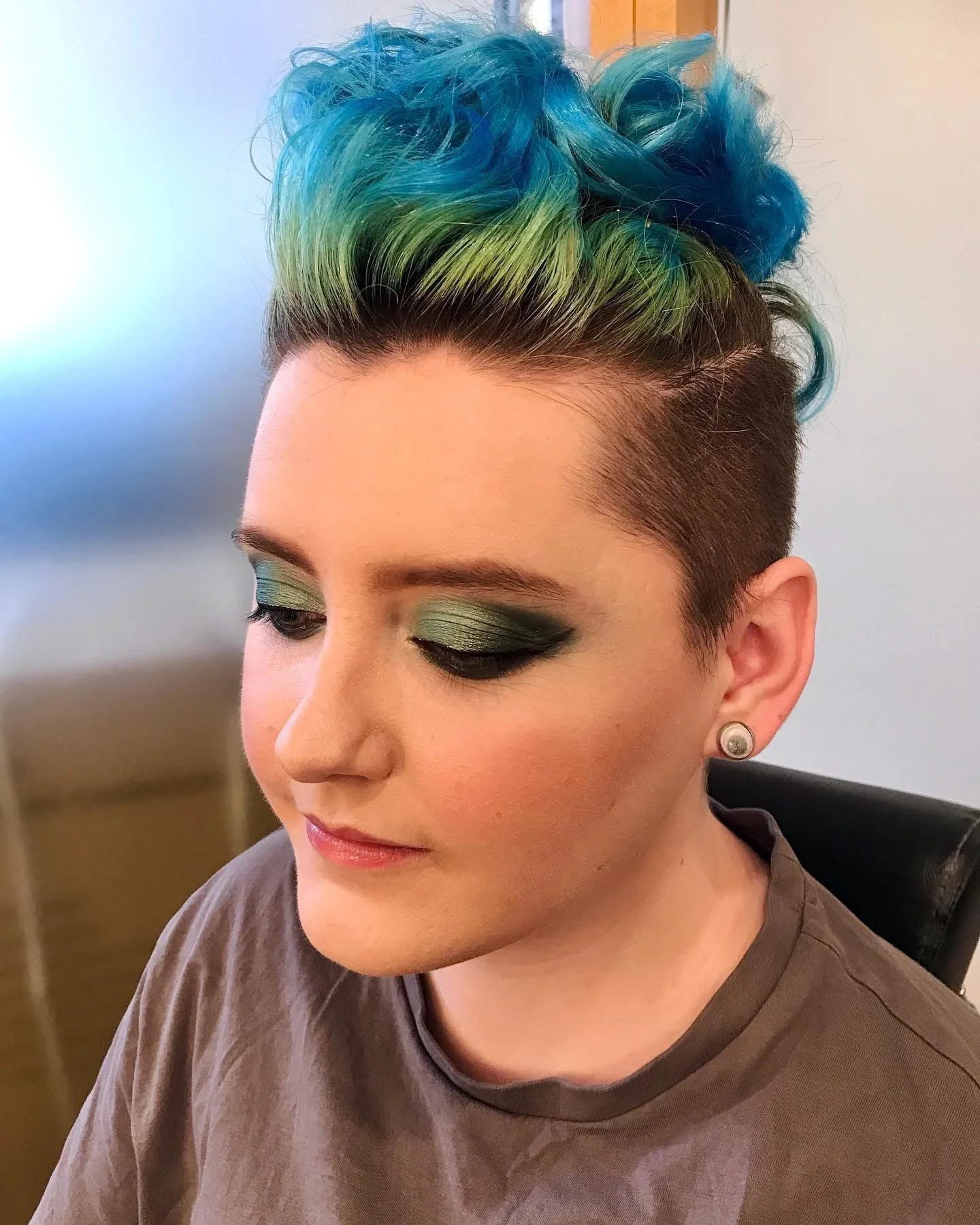 Person with short, rainbow-colored hair styled in curls, wearing green eyeshadow and earrings.