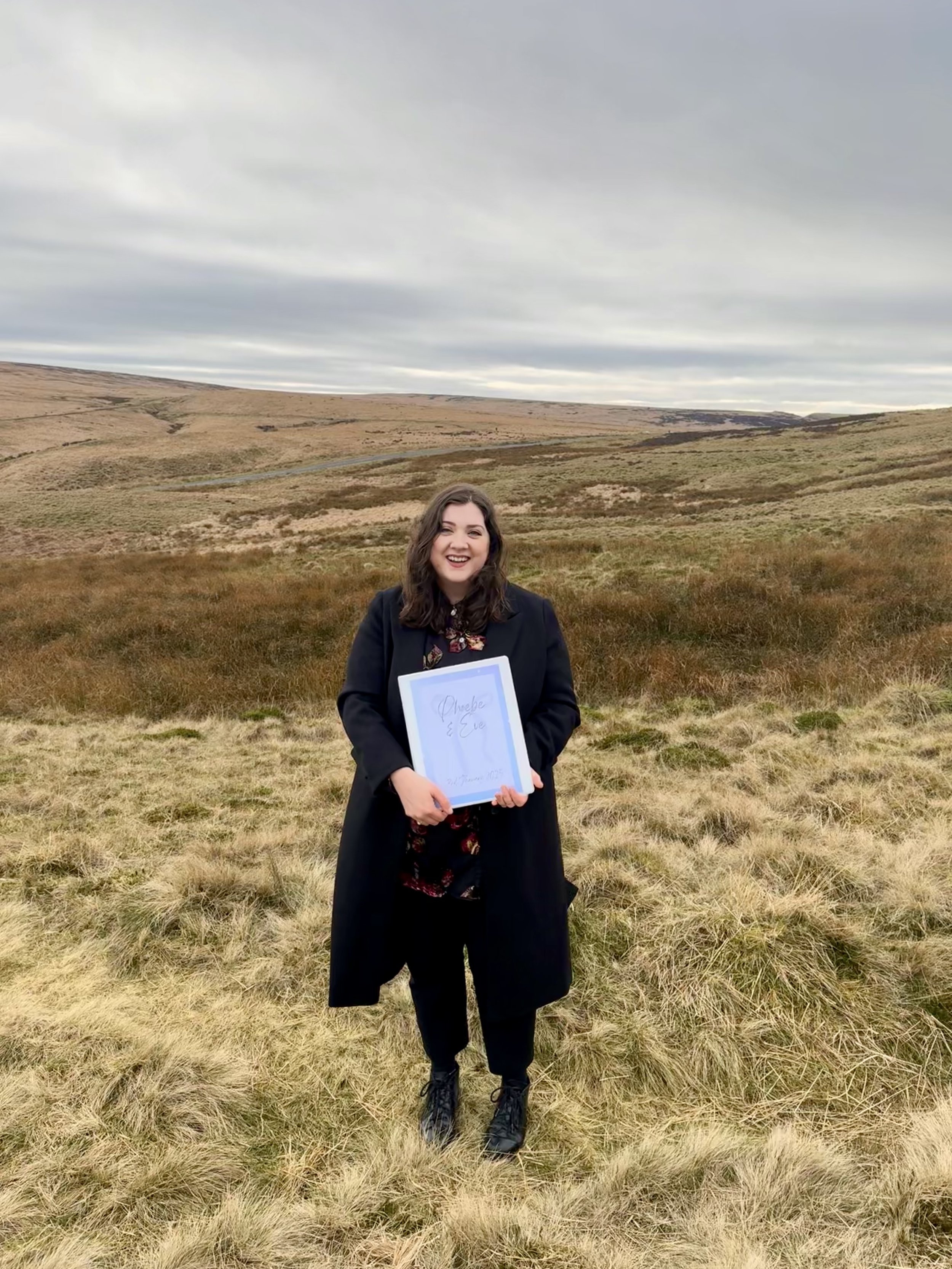 A woman standing outdoors on a grassy field, holding a framed certificate, smiling, with rolling hills and a cloudy sky in the background.