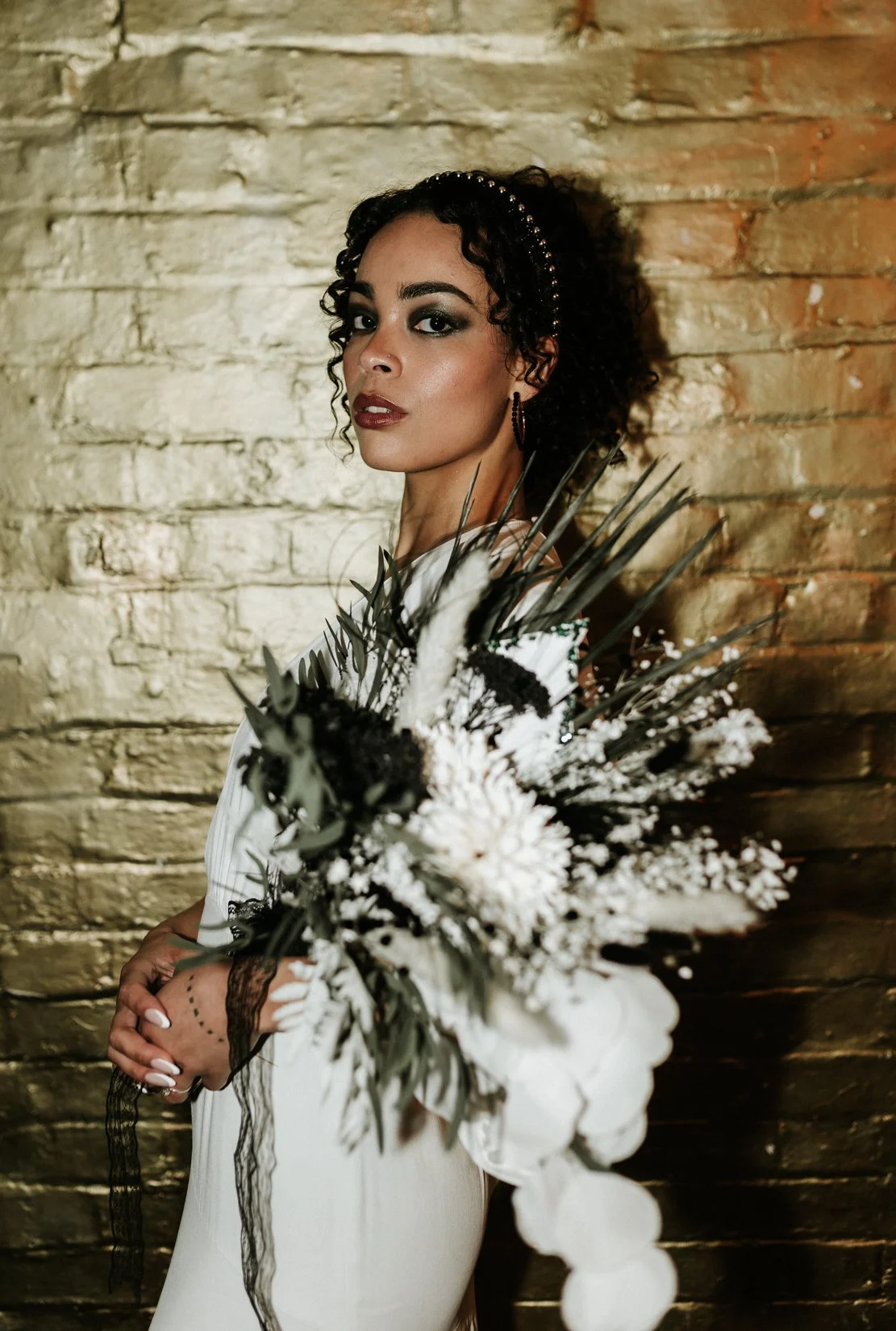 A woman with short curly black hair and dark makeup holding a bouquet of white flowers against a brick wall.