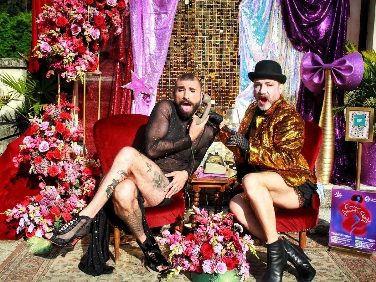Two men dressed in flamboyant clothing, sitting on red velvet chairs surrounded by flowers and colorful curtains, holding phones and singing or speaking.