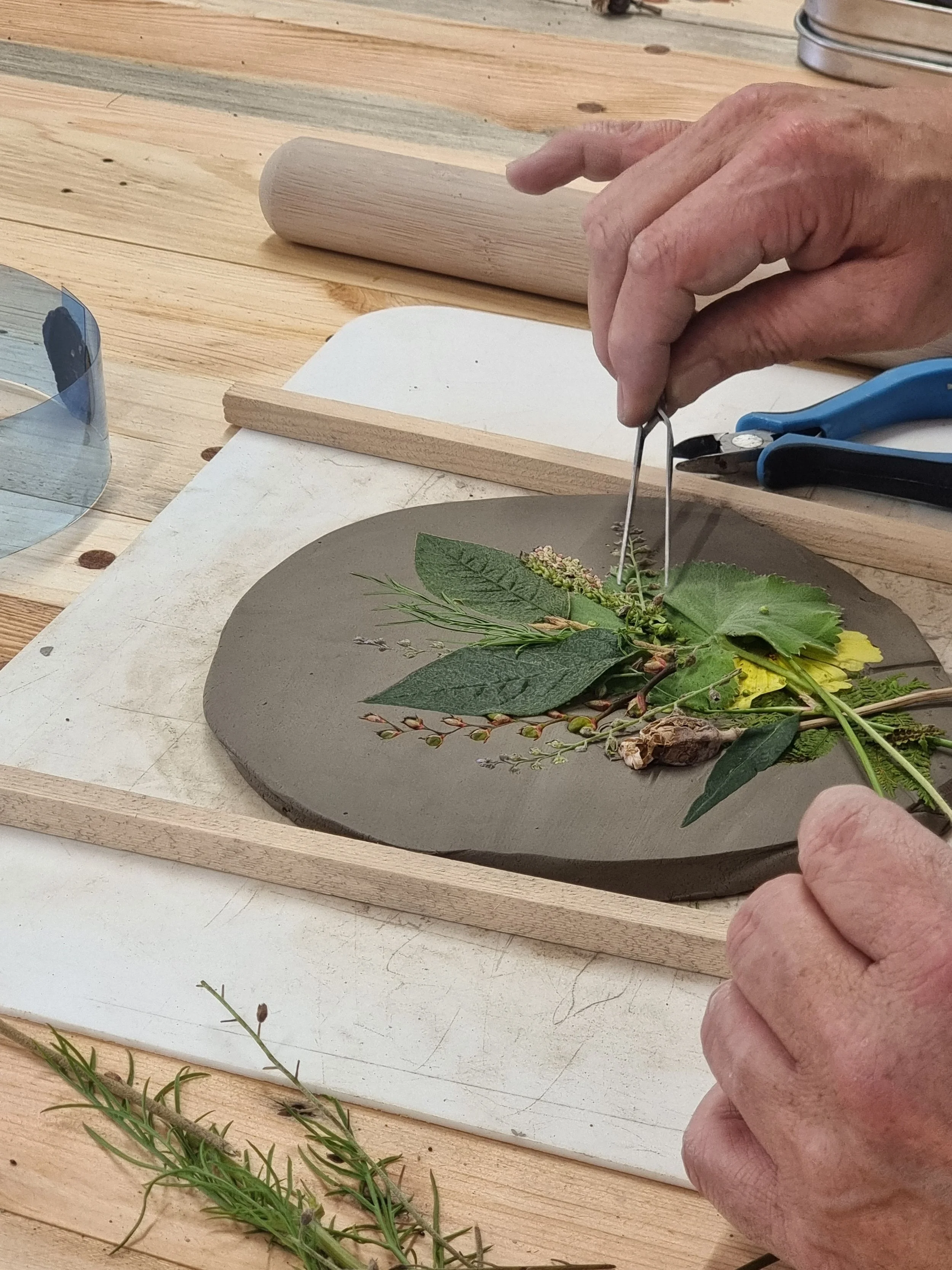 A person is placing a small twig with leaves and flowers onto a circular gray clay or ceramic piece on a wooden table, possibly creating or assembling a botanical artwork or decoration.
