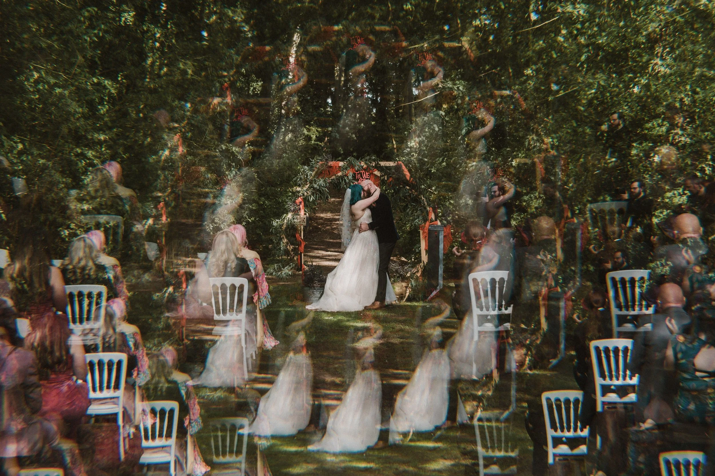 A wedding ceremony taking place outdoors in a forested area with a couple kissing under an arch. Guests are seated on white chairs and multiple exposure effects create ghostly images of the scene.