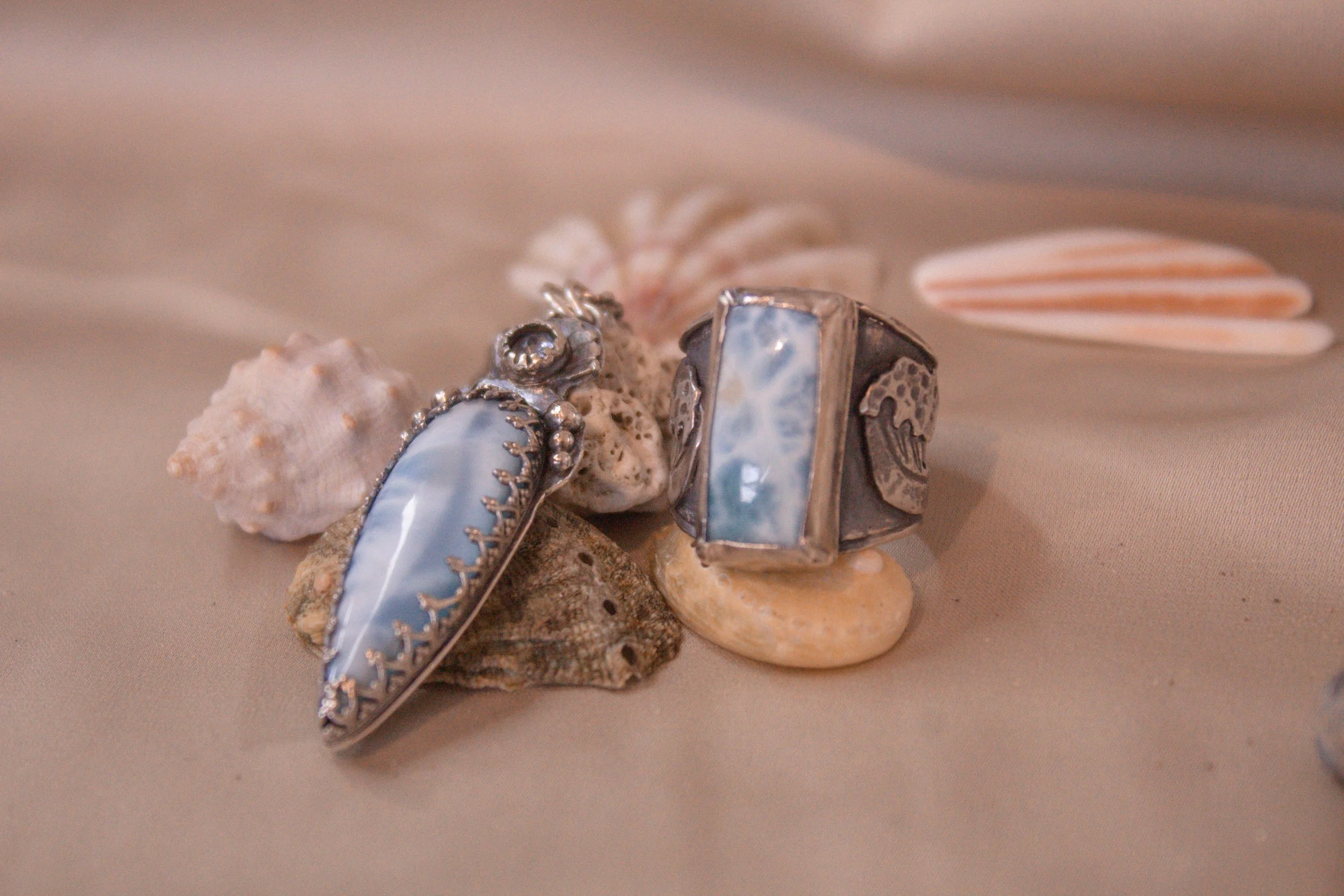 Two silver rings with ocean-blue stones, placed among seashells on a sandy surface.