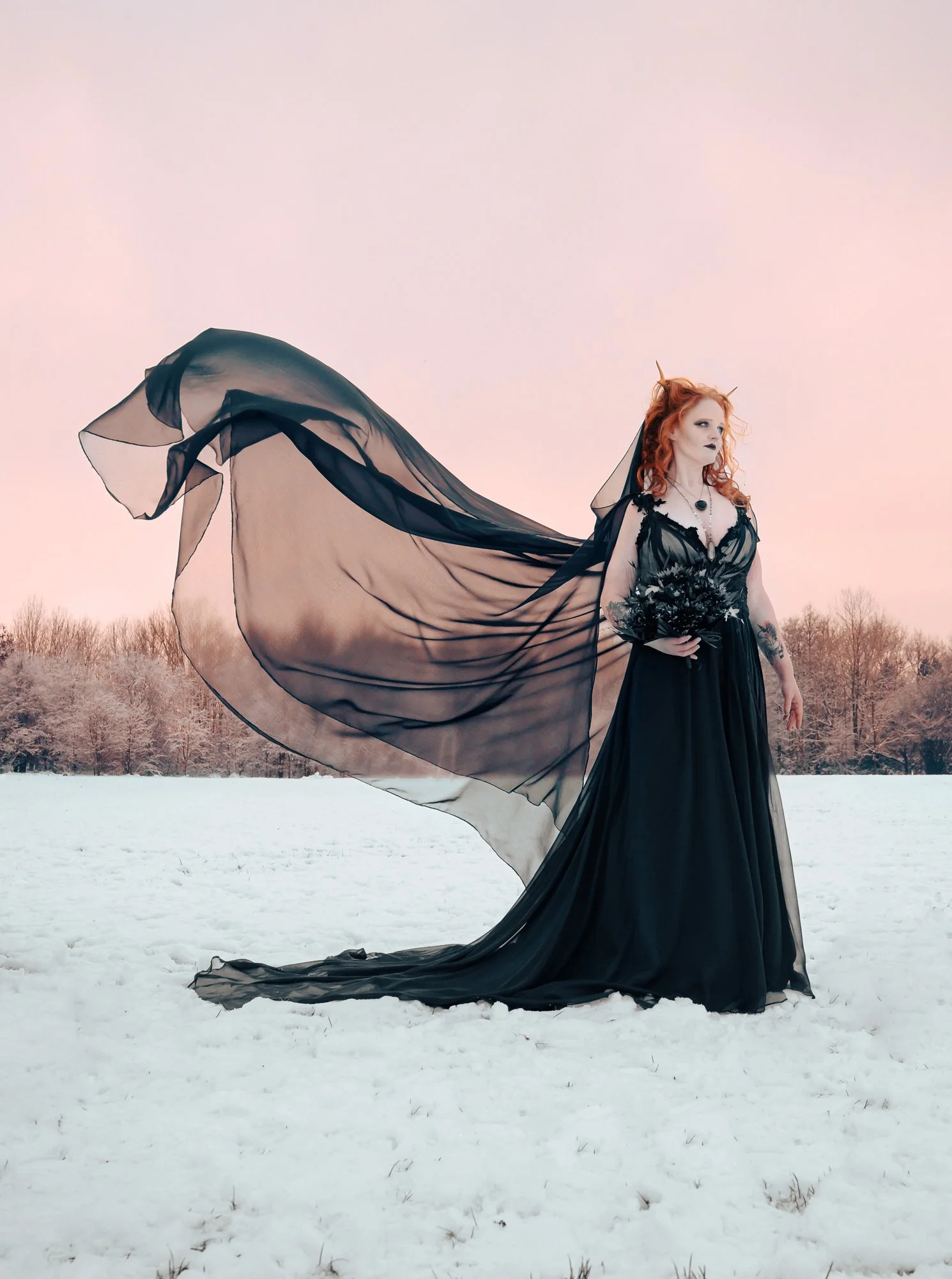 A woman with red hair dressed in a black gothic gown with a flowing sheer cape, standing in a snow-covered field with bare trees in the background during sunset.