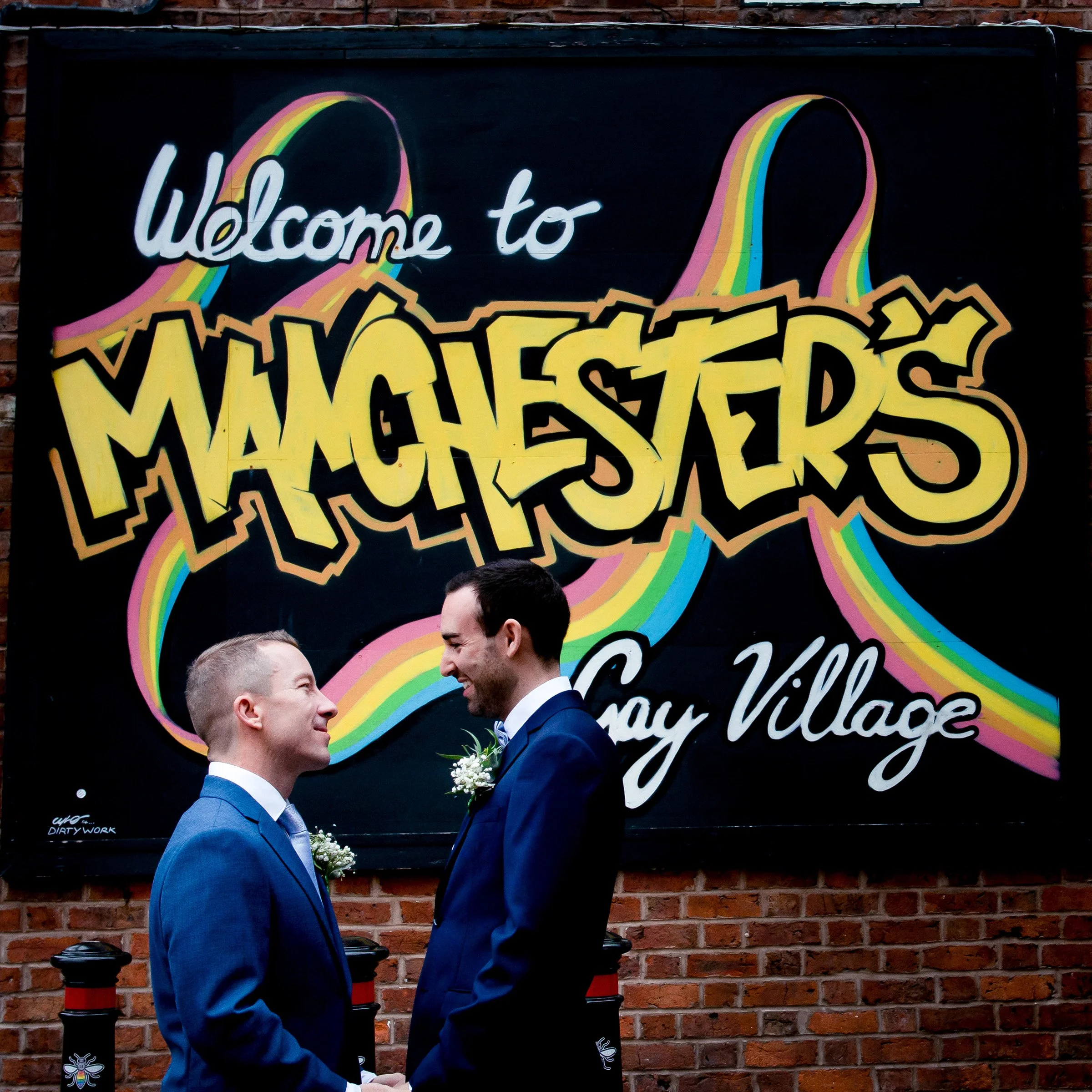 Two men in suits, one with blond hair and the other with dark hair, smiling and holding hands in front of a colorful mural that reads 'Welcome to Manchester, Gay Village' on a brick wall.