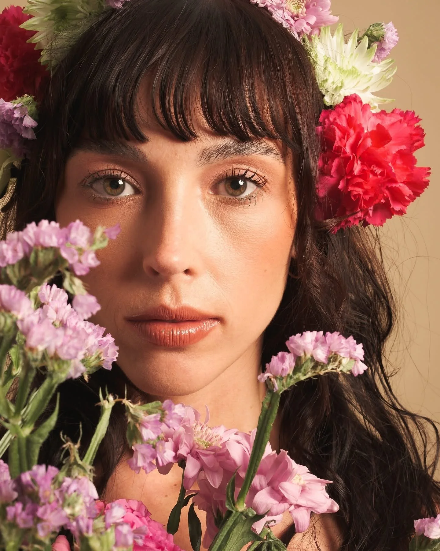 Close-up of a woman with light skin and hazel eyes, surrounded by pink, purple, and white flowers, with dark wavy hair and bangs.