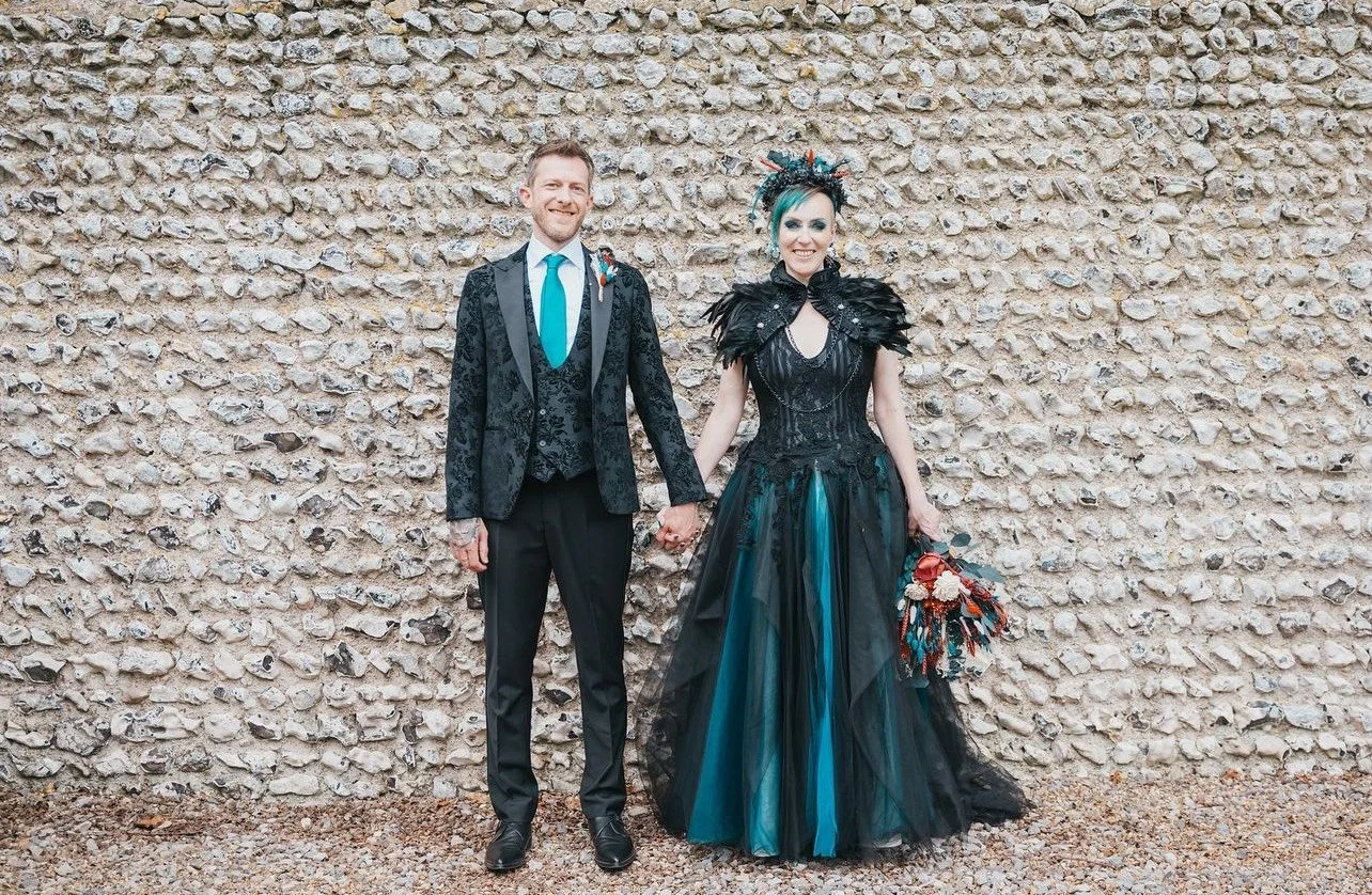 A couple dressed in wedding attire, holding hands and standing in front of a textured stone wall. The man wears a black suit with a patterned blazer, teal tie, and boutonniere. The woman is in a black and teal gown with a dramatic feathered and flora