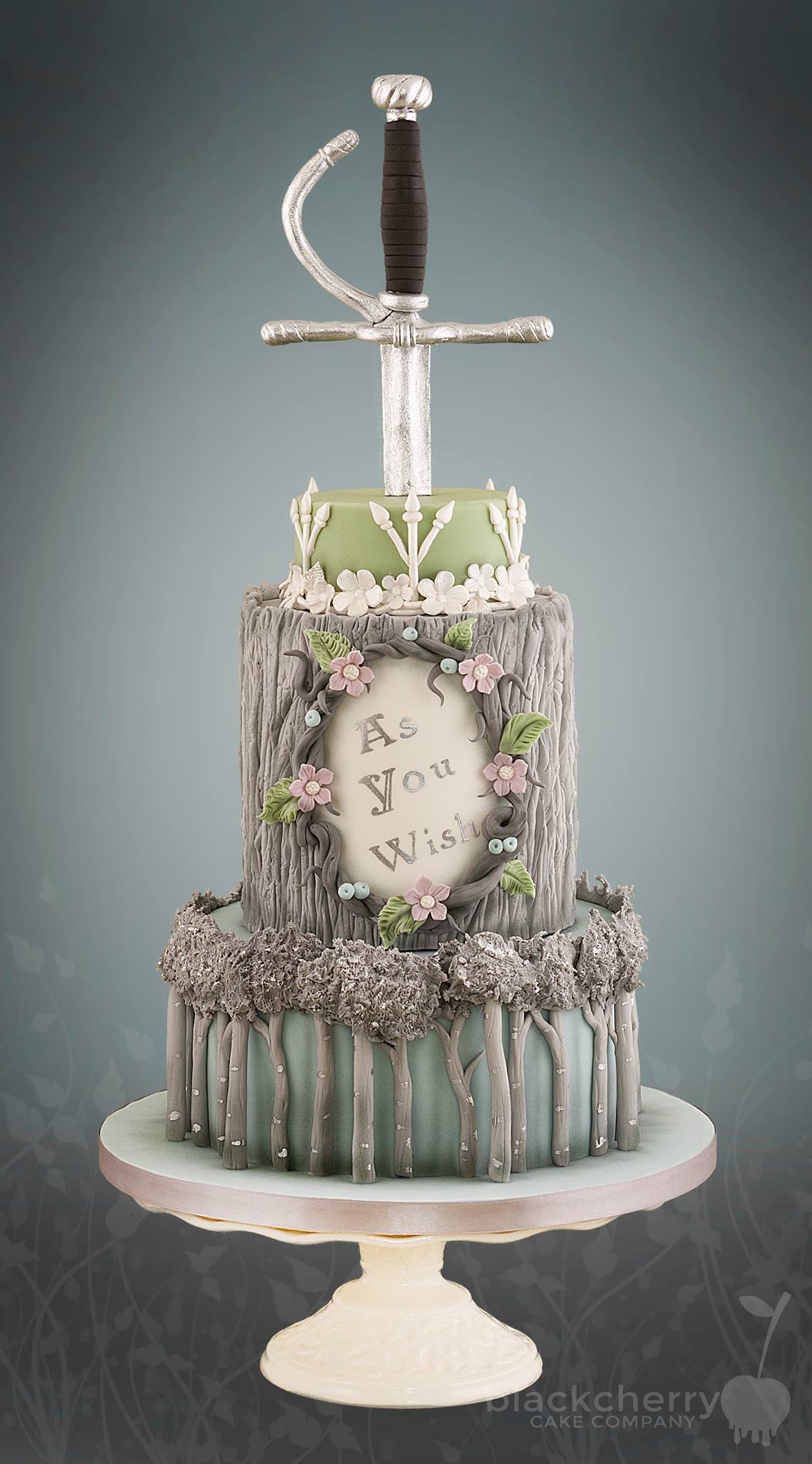 Three-tiered cake with a sword on top, decorated with flowers and leaves, and the words "As You Wish" inscribed on the middle tier.