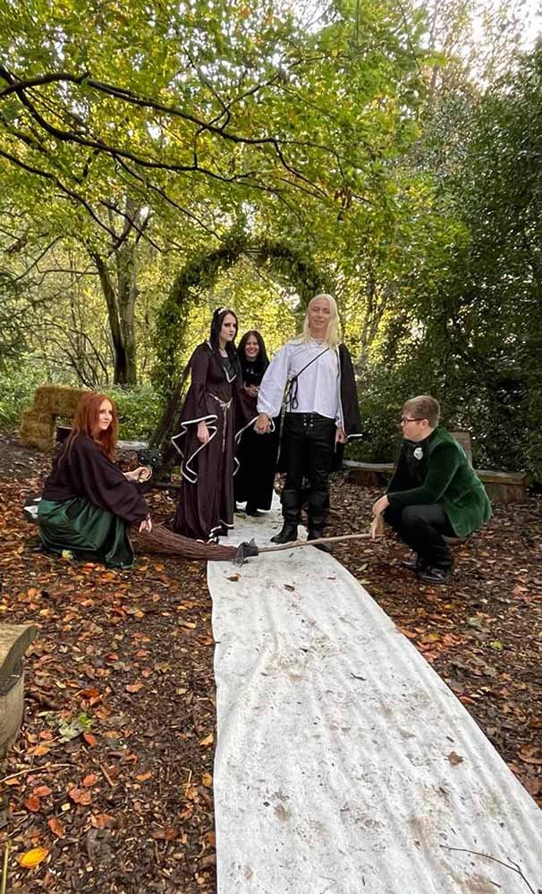 People dressed in medieval costumes standing on a white cloth in a wooded area with a tree arch in the background.