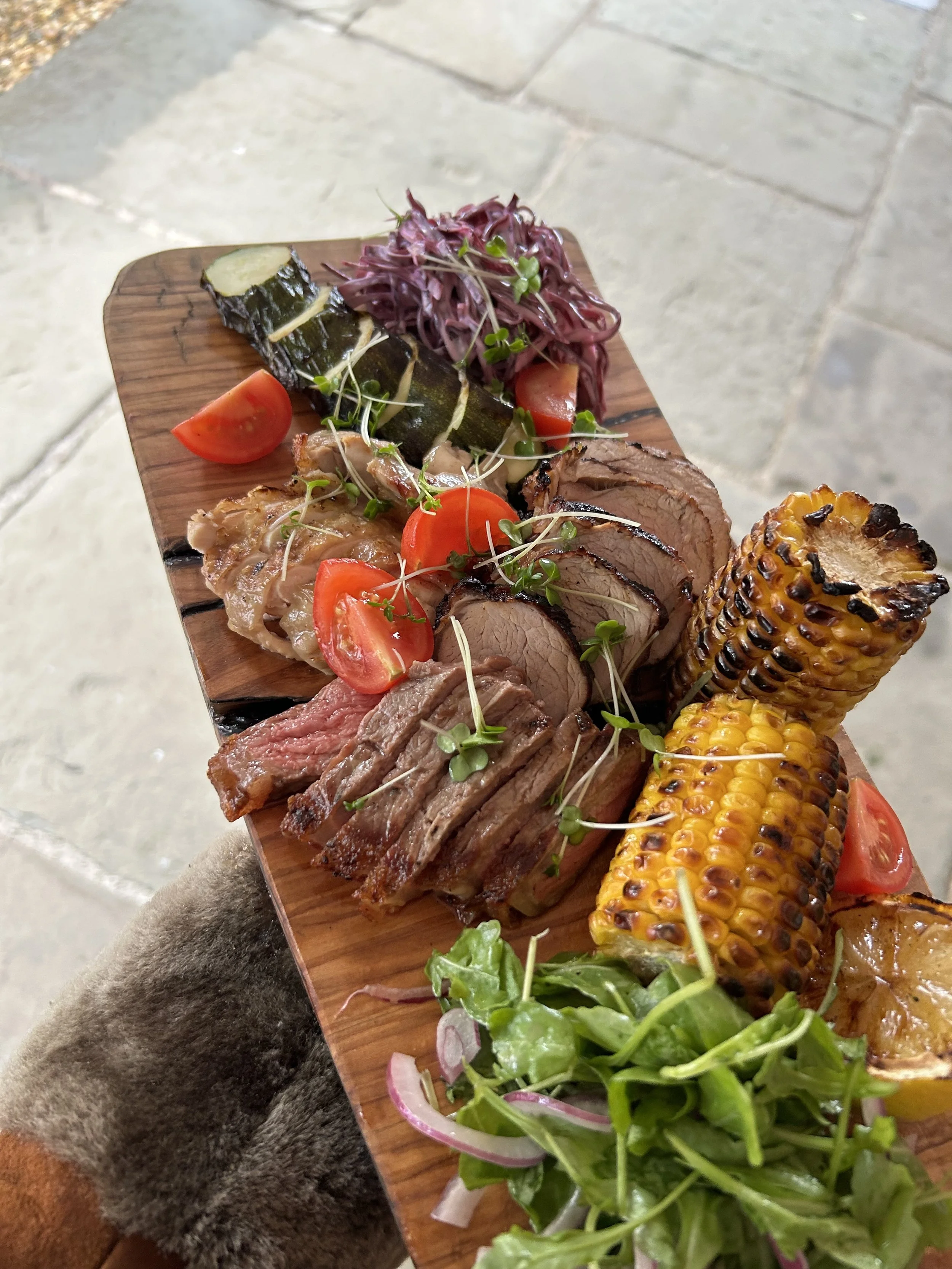A wooden platter with grilled corn, sliced steak, roasted vegetables, cherry tomatoes, fresh greens, purple salad, and microgreens.