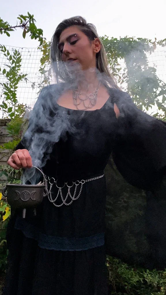 Person wearing black outfit with chains, holding a smoking cauldron outdoors with greenery in the background.